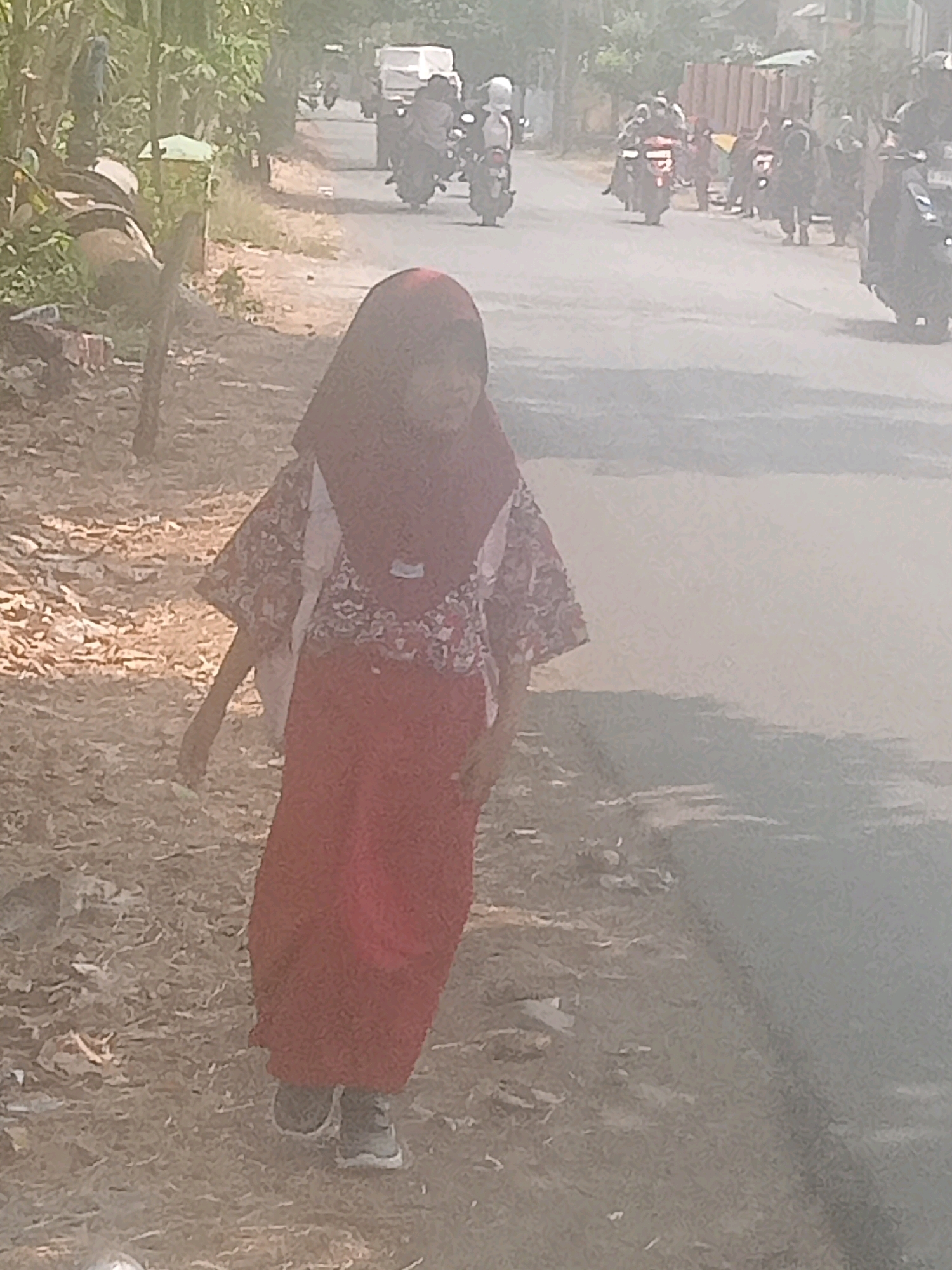 In Lebakgowah, Indonesia, a young girl wearing a red hijab and a patterned top with a red skirt walks on the side of a dusty road. She is wearing grey sneakers. In the background, several motorcycles are visible on the road, some with riders. The environment appears to be a rural or semi-urban area with vegetation lining the road and a hazy atmosphere.