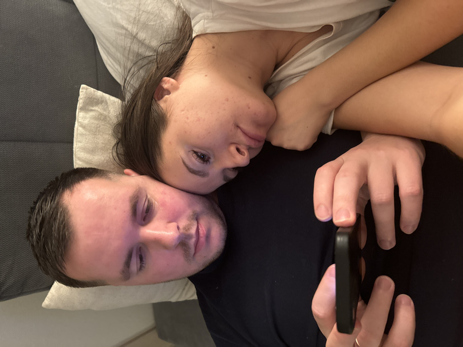 A man and a woman are lying down next to each other. The woman's head rests on the man's shoulder. The man is holding a black mobile phone. The woman is fair-skinned, with dark hair and visible skin texture. The man has short, dark hair. They appear to be on a couch or bed, with pillows visible behind them. The overall lighting is soft and even.