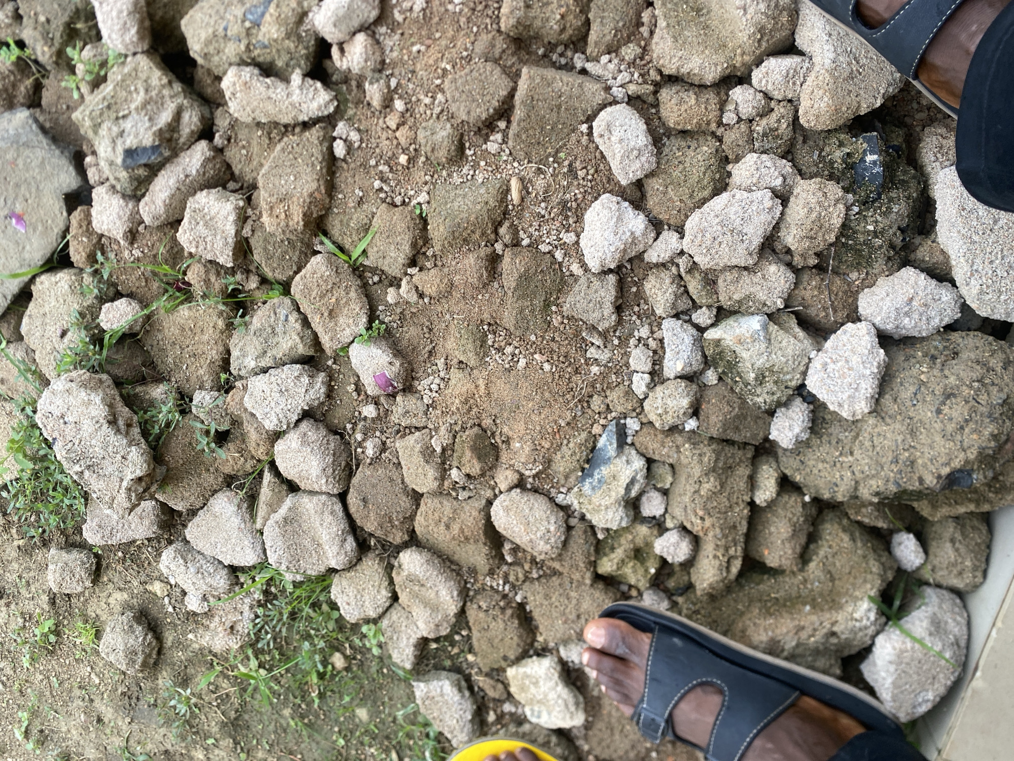 An overhead view shows a person's feet and legs wearing black sandals and a dark garment, placed on a pile of rough, gray and brown rocks and dirt. Small green weeds are scattered amongst the rocks. The scene is outdoors, possibly a construction or demolition site in Dumnga, Nigeria. The lighting suggests it is daytime.