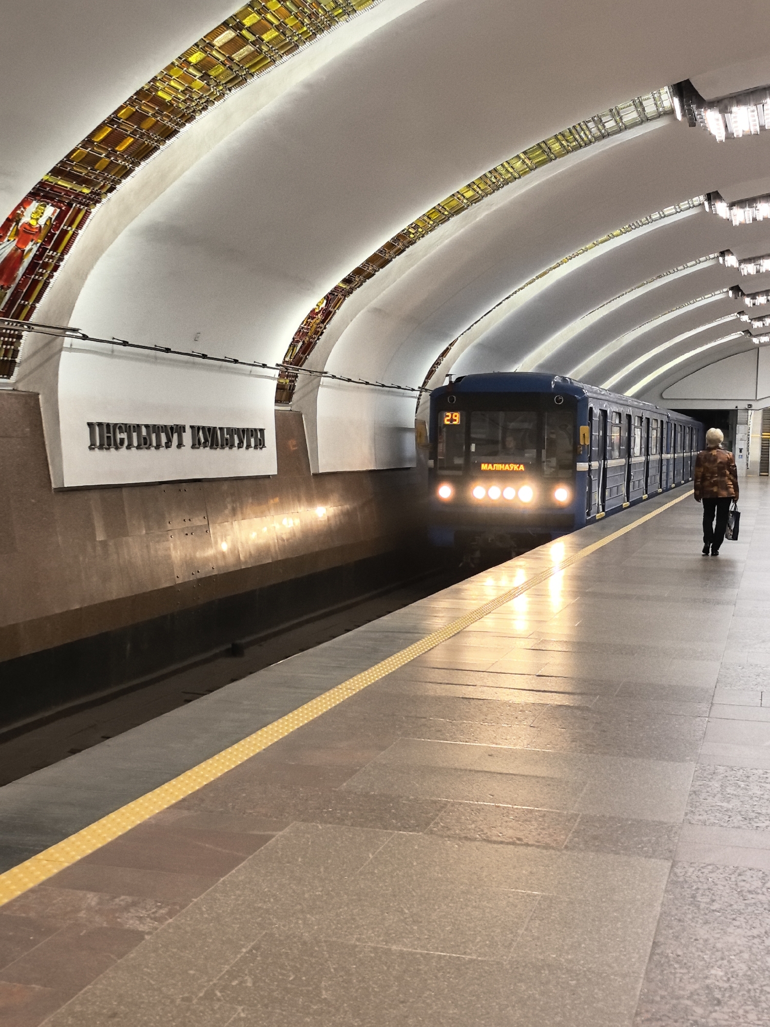 A blue subway train is stopped at the Institut Kultury station in Minsk, Belarus. The station has a vaulted ceiling decorated with stained glass mosaics and modern light fixtures. The platform is paved with stone tiles and marked with a yellow safety line. A woman in a patterned jacket and black pants is walking away from the camera down the platform, carrying a black bag. The train's headlights are on, illuminating the platform. The station name "Institut Kultury" is visible on a white sign above a dark, textured wall.