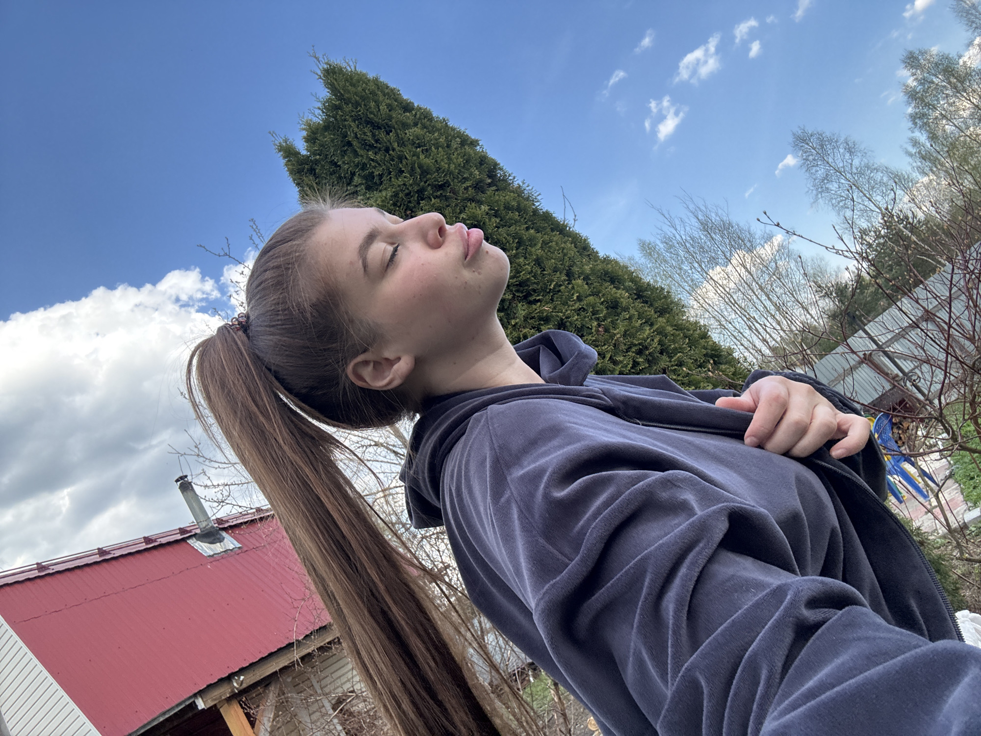 A young woman stands outdoors, her head tilted back with eyes closed, appearing to bask in the sun. She has long brown hair pulled back into a ponytail. She wears a dark blue or gray zip-up jacket. Behind her is a blue sky with scattered clouds and a tall green evergreen tree. In the background, there is also a red roof of a building and some bare trees. The location is not identifiable as Мещёры, Russia based on the image.