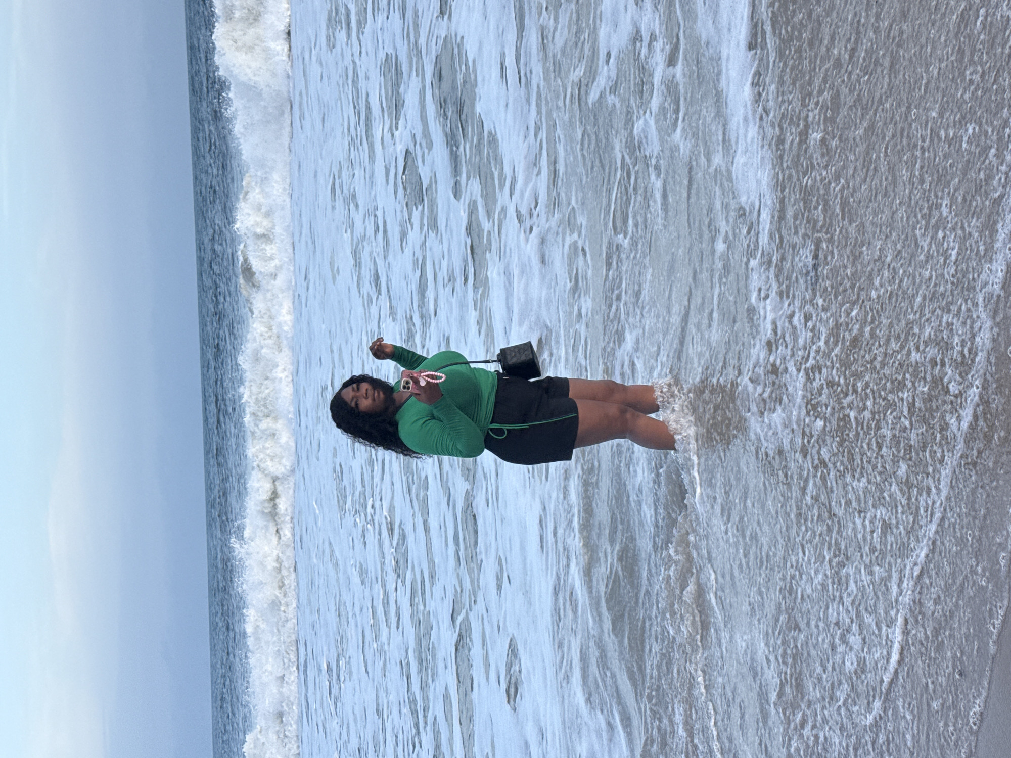 A person stands in the surf at a beach in Mopo Onijebu, Nigeria. The individual is wearing a long-sleeved green top and black shorts. A small black purse is slung over their shoulder. They are holding a cell phone up in their hand. The ocean water surrounds their legs, and waves are breaking in the background. The sky is overcast. The sand is light in color.