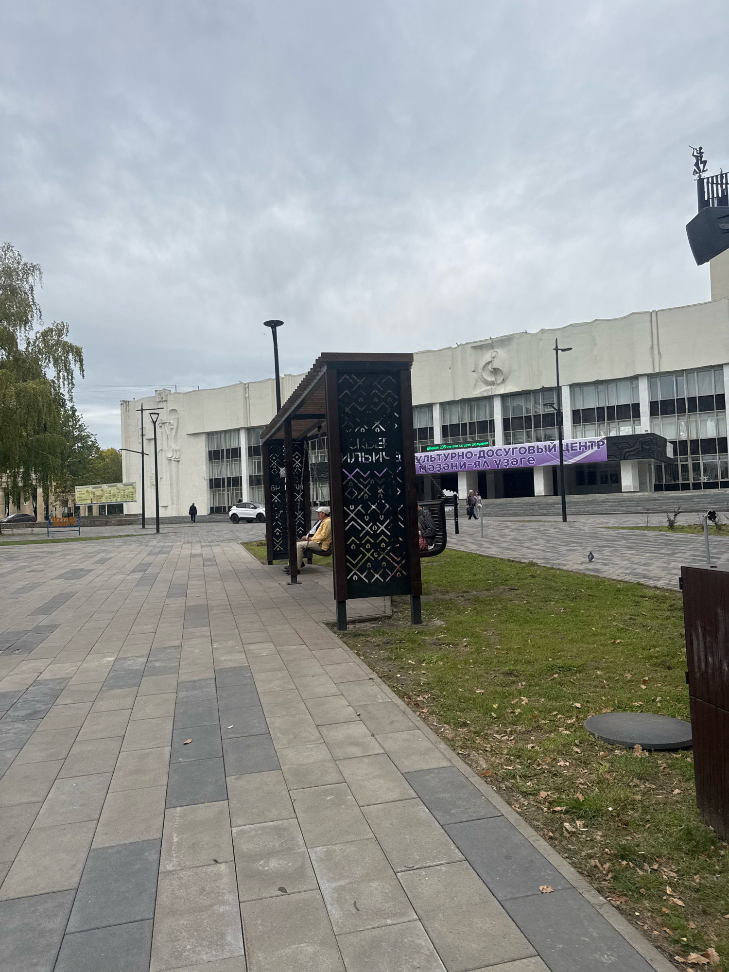 The image depicts a paved walkway in front of a large, white building in Ufa, Russia. The building has a prominent banner across its facade that reads "КУЛЬТУРНО-ДОСУГОВЫЙ ЦЕНТР МЭҲЭМЭН-ЯЛ ҮЗӘГЕ" (Cultural and Leisure Center Mehman-Yaluge). To the right of the walkway, there is a wooden structure with a bench swing, occupied by two people. A third person is walking away from the viewer towards the building. The sky is overcast. The paved area is made of large, rectangular stones in shades of grey and beige. Grass and fallen leaves are visible on the right side of the walkway.