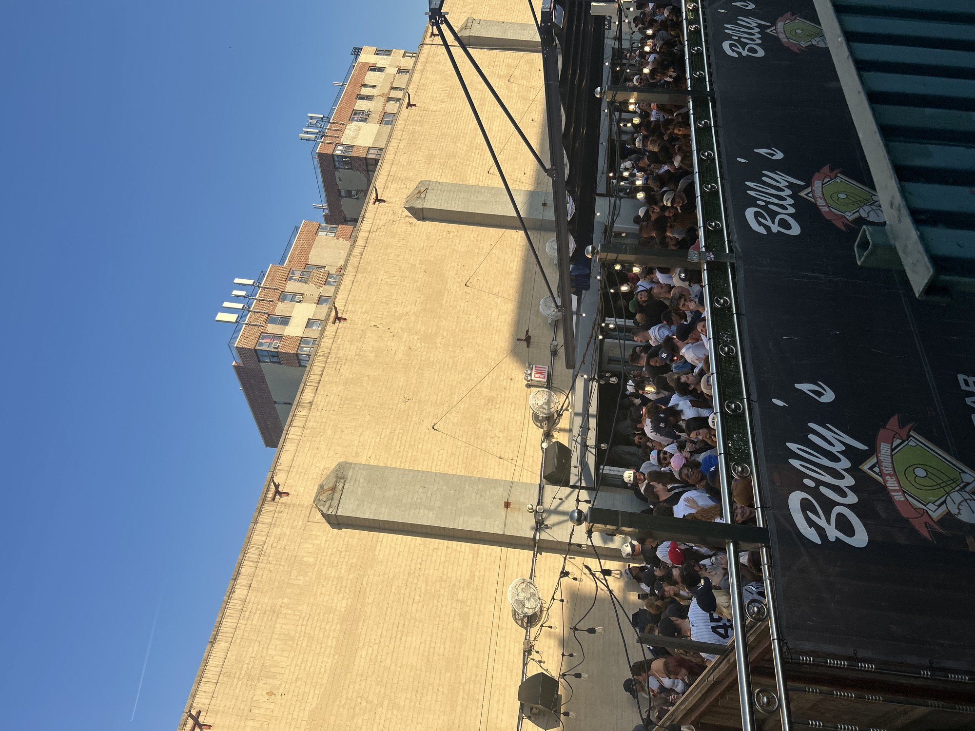 The image shows a view from a crowded outdoor bar, presumably in New York City, United States of America, given the large number of people wearing New York Yankees apparel. The bar is named "Billy's at the stadium" as evidenced by the signage. The crowd is densely packed on a balcony or raised area, suggesting a popular gathering place. A building with a brick facade is visible in the background, and a clear blue sky is seen above. The bar appears to be situated close to a stadium. There are light bulbs hanging above the patrons. Also visible are fans and speakers.