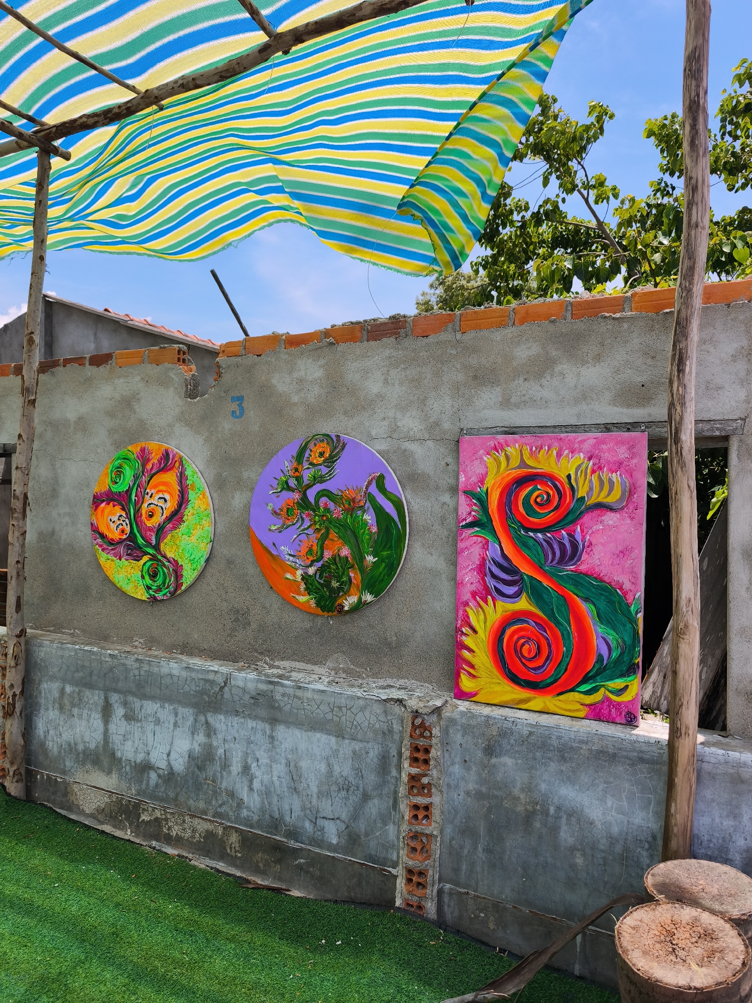 An outdoor scene in Vĩnh Lương, Vietnam features a concrete wall adorned with three colorful, abstract paintings. Two of the paintings are circular, one predominantly green and orange with swirling floral-like patterns, and the other purple with similar organic designs and orange accents. The third painting is a rectangular canvas with a vibrant pink background, depicting a stylized, swirling creature in shades of orange, green, and yellow. Above the wall, a striped awning made of green, yellow, and blue fabric is supported by wooden poles. In the foreground, artificial green turf covers the ground, and two tree stump-like objects are visible to the right. The wall itself is weathered, with some exposed brickwork and a number "3" painted in blue. The background shows a clear blue sky and some green foliage from trees.