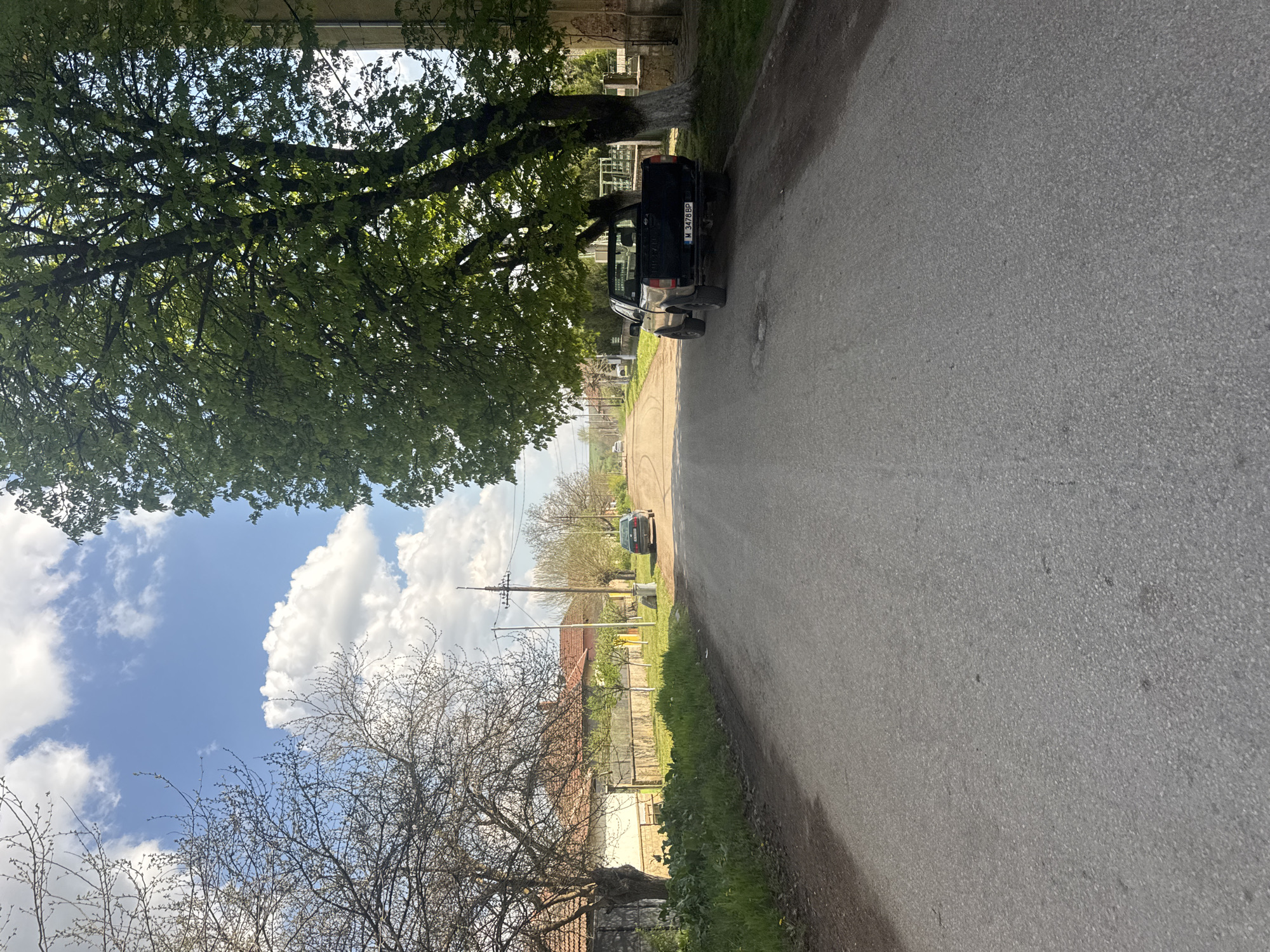 A daytime street scene in Lehchevo, Bulgaria features an asphalt road extending into the distance. A black pickup truck is parked on the right side of the road, partially under a large, leafy tree. On the left, another tree with bare branches is visible against a backdrop of a blue sky with cumulus clouds. In the distance, houses and vegetation line the road. A utility pole is also visible.