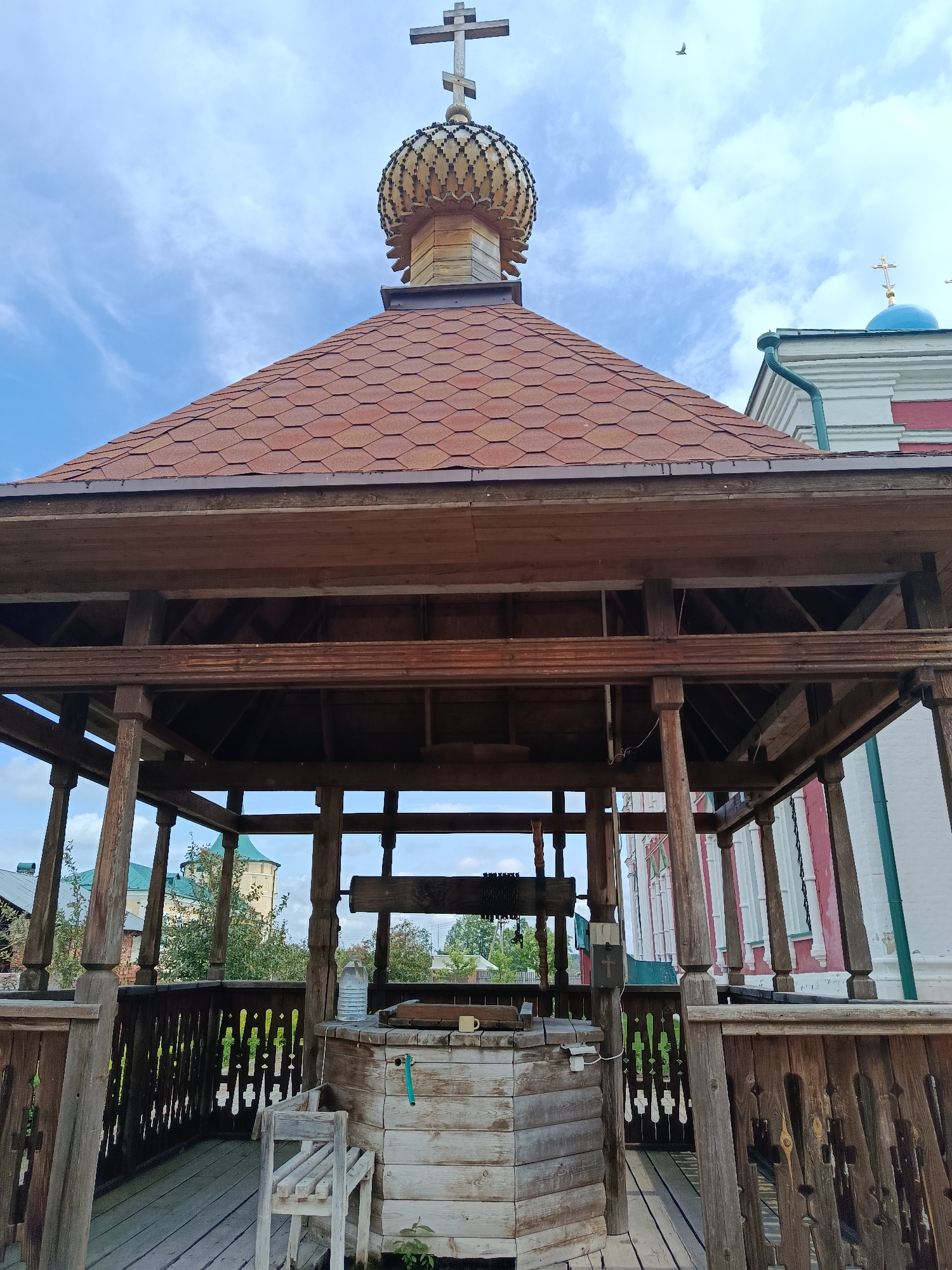 The photograph captures an outdoor scene featuring a wooden well structure in the foreground, with a domed roof topped by a cross. The well itself is made of light-colored wood planks. A wooden bench is positioned to the left of the well. The well structure is surrounded by a wooden deck and railing.
In the background, a building with a red and white facade and a green roof is visible, as well as other structures and trees. The sky is blue with scattered clouds.
The image is identified as being taken in Radovitsy, Russia, likely at a holy site or historical location given the presence of religious architecture. The photograph is taken during daylight hours. There are no people visible in the image. The overall impression is one of a traditional and possibly sacred site.