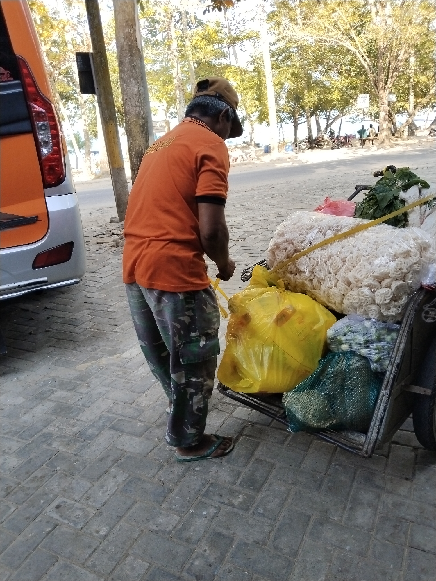 A man in an orange t-shirt and camouflage pants stands next to a cart laden with goods, possibly near the beach in Prigi, Indonesia. The cart contains large bags of white, coiled snacks, a bright yellow plastic bag, and a green mesh bag filled with produce. A portion of an orange and silver van is visible on the left side of the frame. The ground is paved with grey interlocking bricks. In the background, trees line a street, and several people are visible further down, some near motorcycles and benches, suggesting a public area or roadside setting. The lighting indicates it is daytime, with sunlight casting shadows.