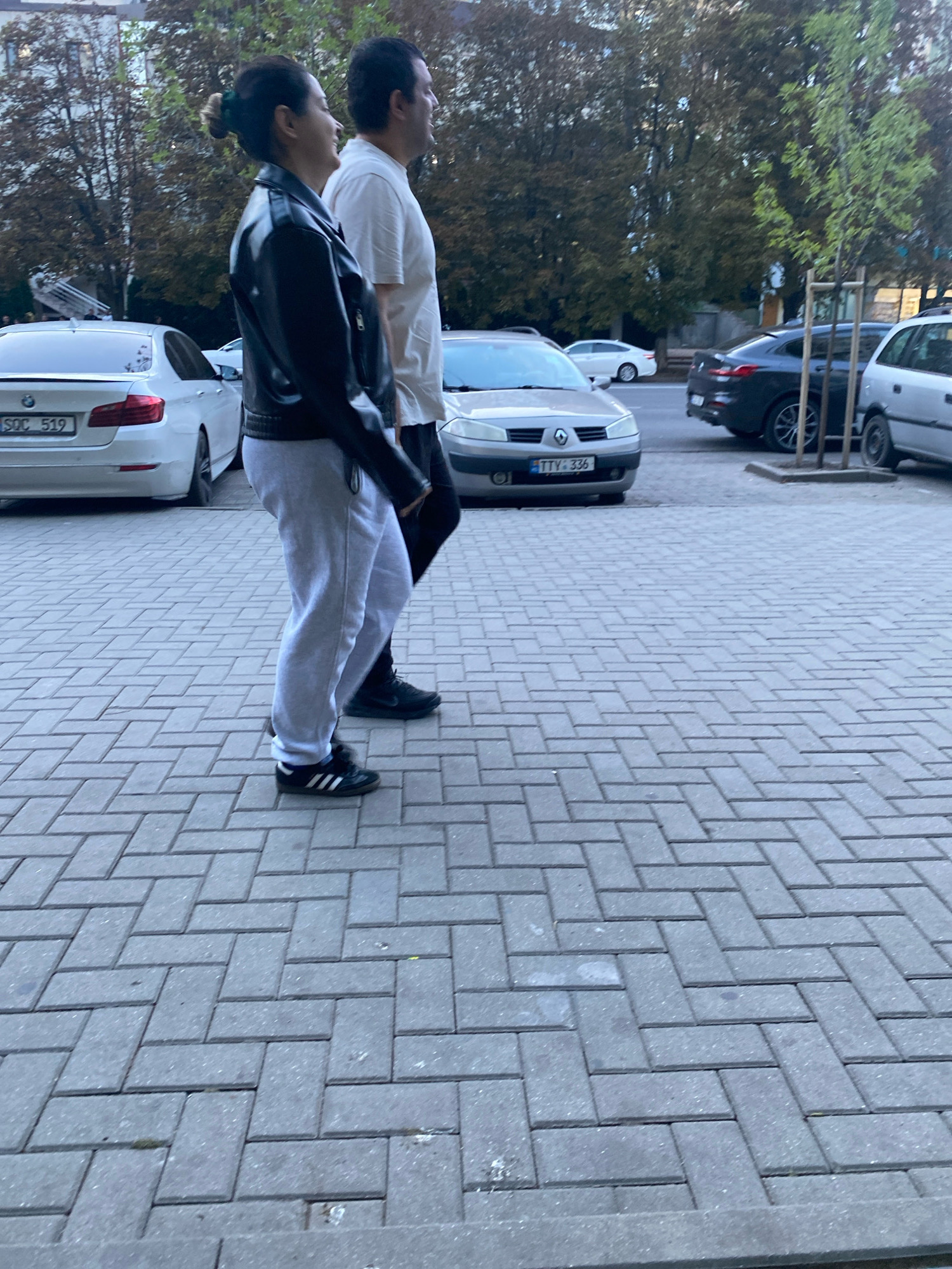 The image shows two individuals, a woman and a man, walking side-by-side on a cobblestone sidewalk. The woman is on the left, wearing a black leather jacket, grey sweatpants, and black Adidas sneakers. Her hair is pulled back in a bun, adorned with a green scrunchie. The man is to her right, wearing a light-colored t-shirt and dark pants. They are walking away from the camera and appear to be moving in the same direction. In the background, several cars are parked on the street, including a white BMW with the license plate "SOC 519" and a dark sedan with the license plate "TTV: 336". Trees with foliage are visible behind the cars and the people. The setting appears to be a street in Chișinău, Moldova, given the context of the provided information. The scene is captured during daylight.