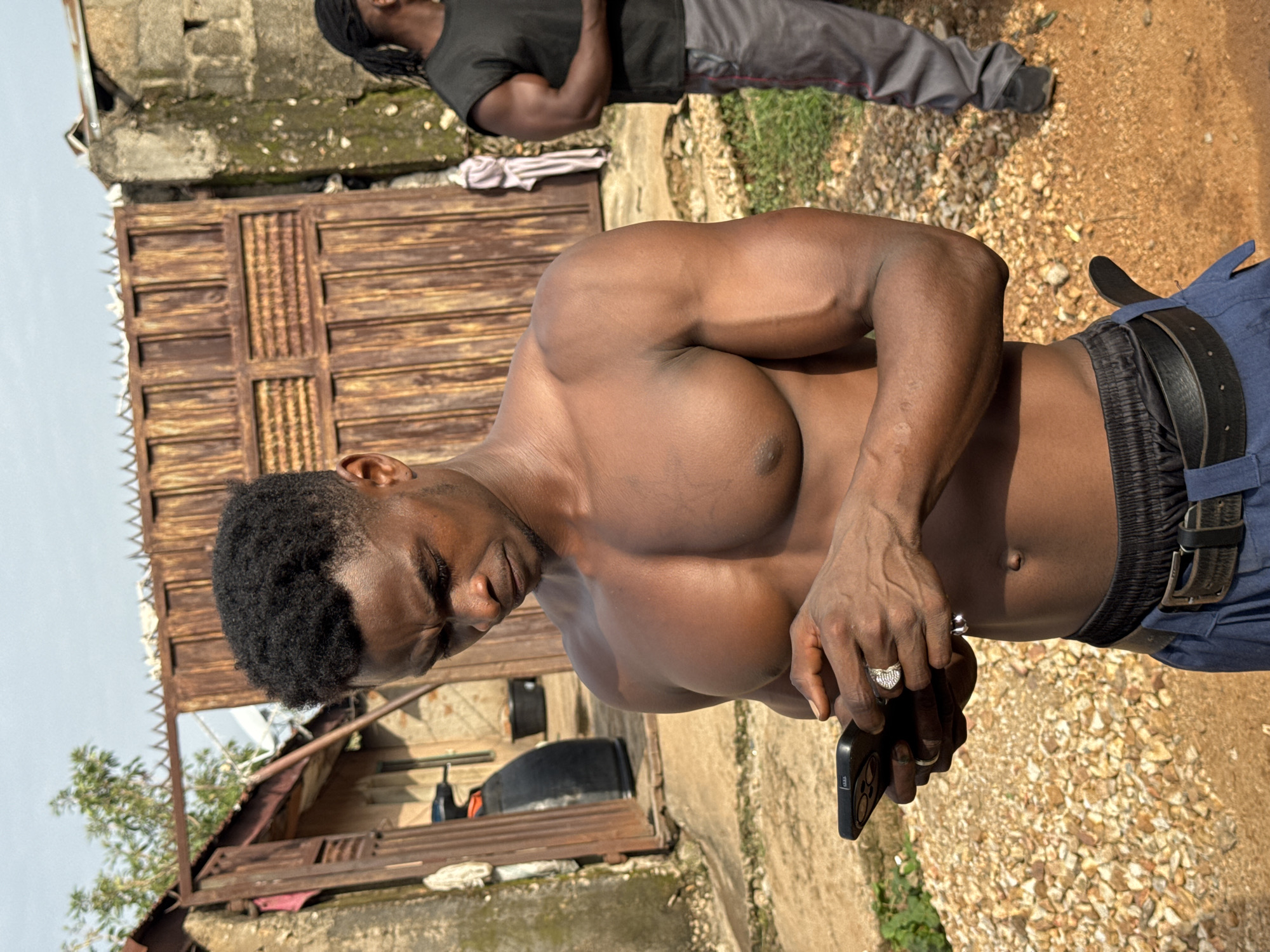 A shirtless man with dark skin and short black hair is standing in an outdoor area, looking down at a smartphone in his hands. He is wearing blue pants with a black belt. His muscles are defined, particularly his chest and arms. To the right, another man with dark skin and braided hair pulled back is visible, wearing a black t-shirt and gray pants. Behind the first man is a rustic wooden gate and a building. The ground is covered with gravel and dirt. The scene appears to be in a somewhat unpaved area, possibly in a less developed part of the city of Akwanga, Nigeria.