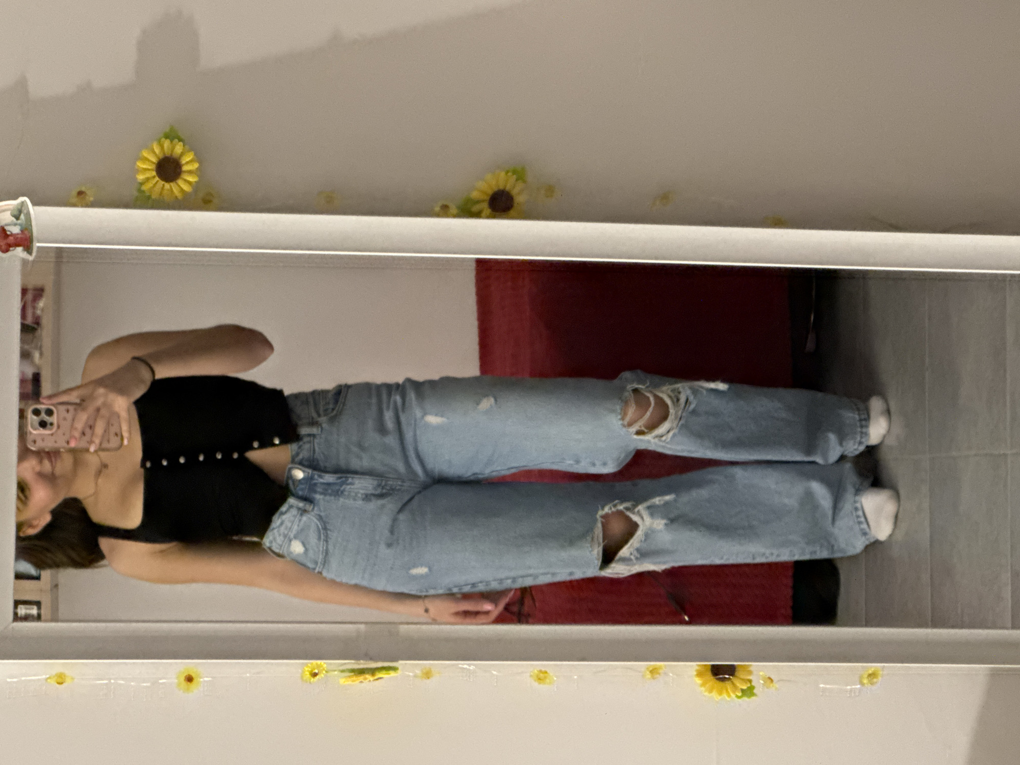 A young person, reflected in a mirror, is taking a selfie. The person is wearing a black top and distressed, light-wash denim jeans. The location appears to be a domestic interior. The mirror is decorated with artificial sunflowers. There is no evidence to suggest that this image was taken in Sofia, Bulgaria.