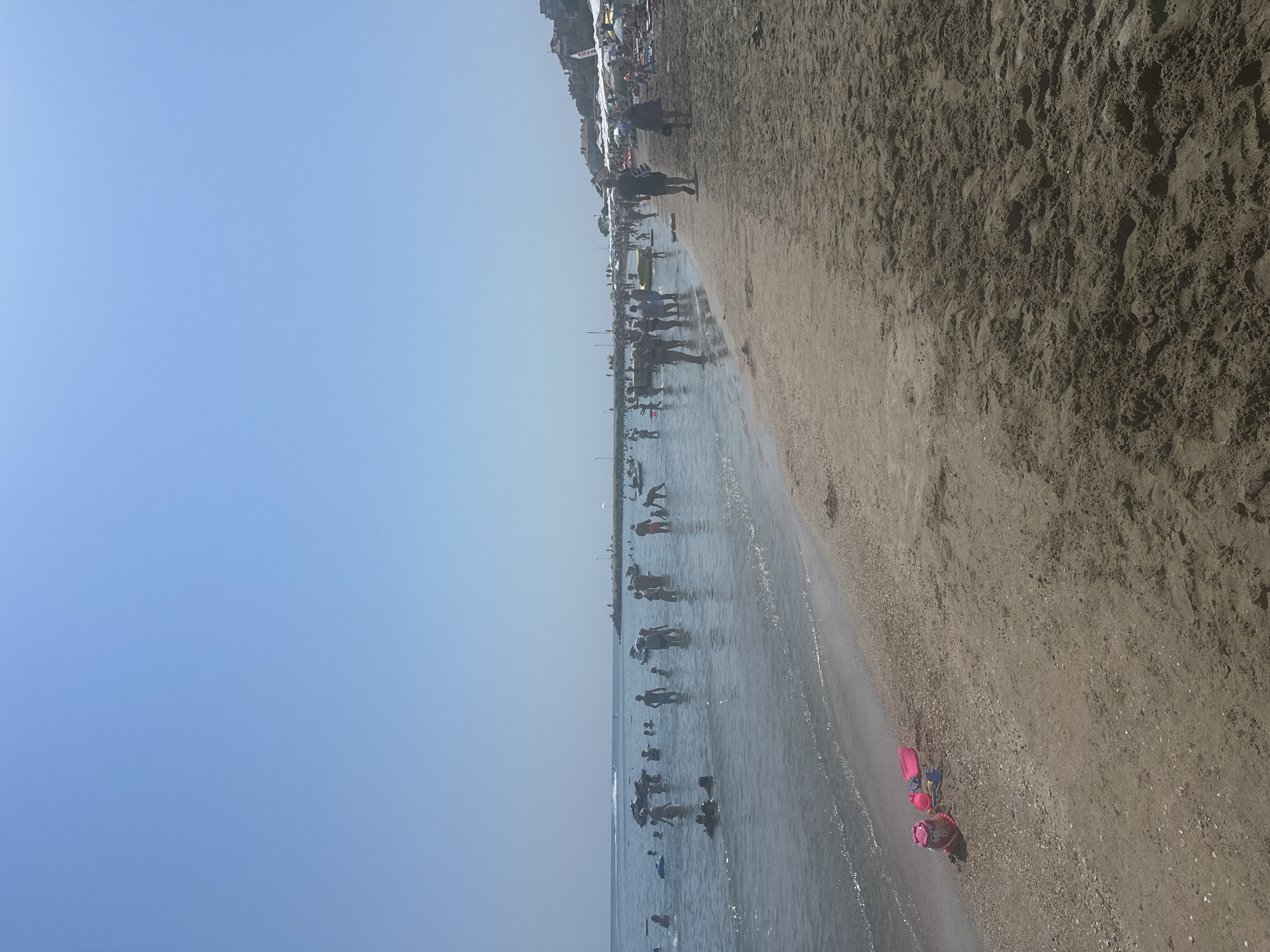 The image depicts a crowded beach scene on a sunny day. The foreground is dominated by sand, with a small collection of pink and blue children's beach toys on the left. The sea occupies the middle ground, with numerous people wading, swimming, and standing in the shallow water. Several individuals are visible in the water, some appearing to be adults and children. In the background, a breakwater extends into the sea, with more people and beach umbrellas visible on the shore beyond. Structures, likely buildings, are nestled on a hill overlooking the beach. The sky is clear and blue. The location is identified as Pomorie, Bulgaria.