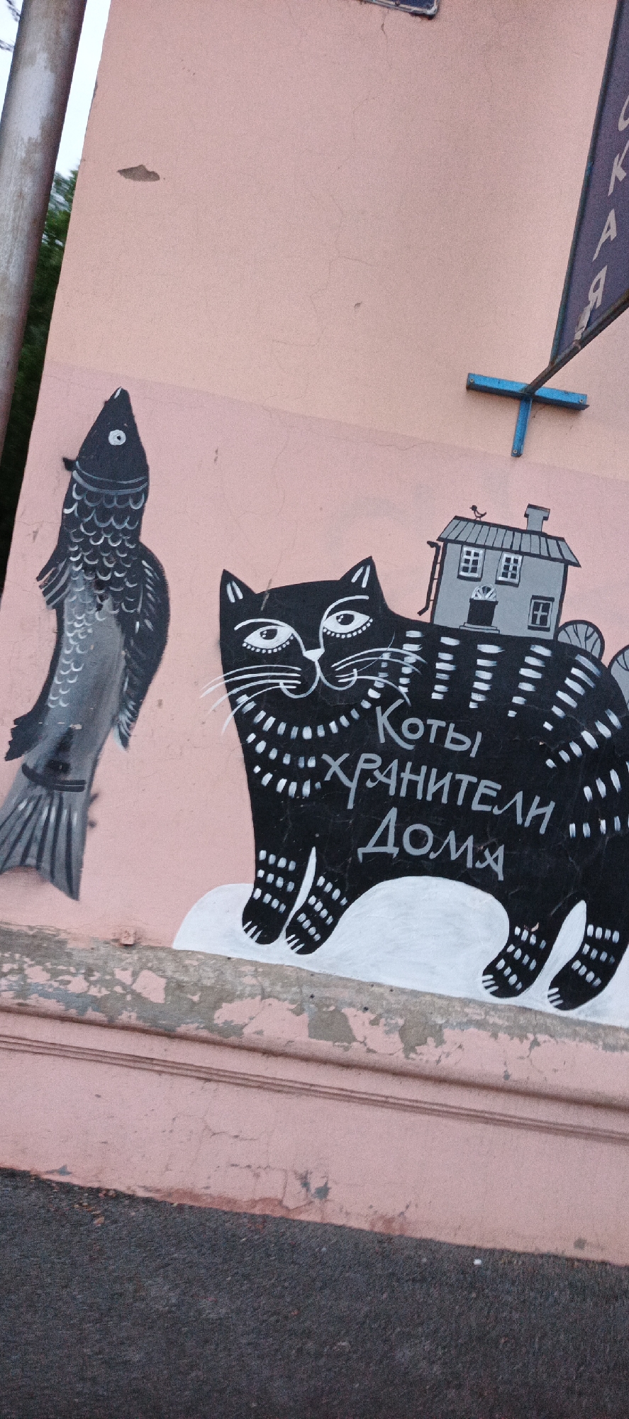 The image captures a section of a pale pink, textured wall with graffiti art. On the left, a black and white stylized fish is depicted vertically. To its right, a large black cat with white markings is painted. The cat is wearing a small grey house on its back, and a tiny bird is perched on the roof of the house. White text in Cyrillic is painted across the cat's body, reading "Коты хранители дома" which translates to "Cats are guardians of the home." The wall shows signs of wear and tear, with peeling paint and visible cracks. In the upper right corner, a blue sign with white Cyrillic lettering is partially visible, appearing to be an outdoor advertisement. The ground in front of the wall is asphalt. The scene appears to be an exterior wall in the city of Votkinsk, Russia, likely on a street or public space.