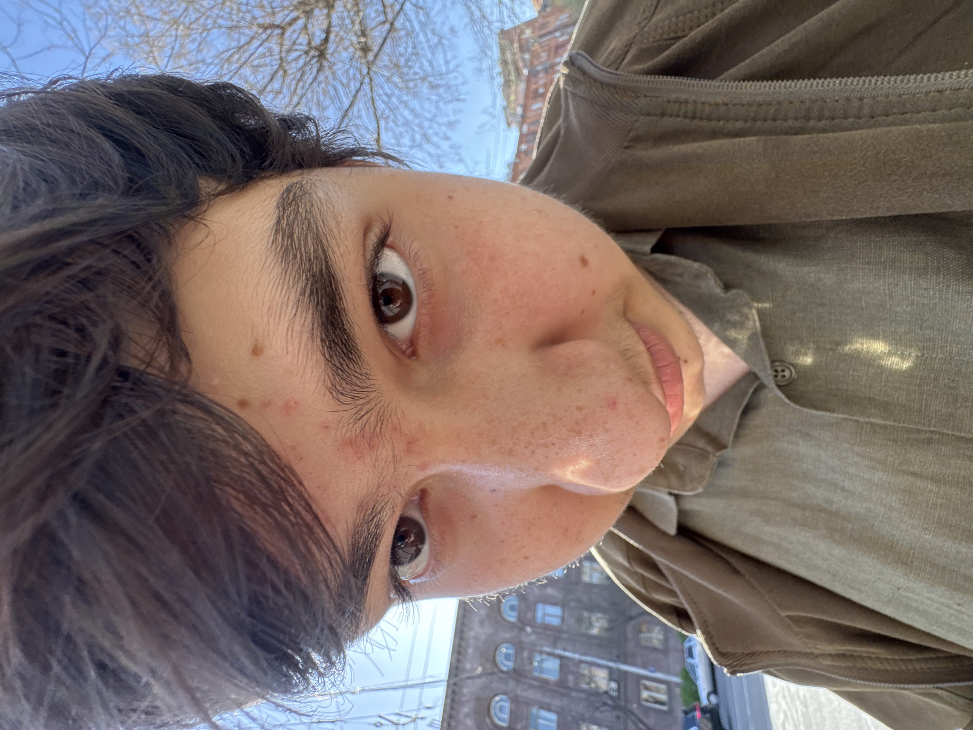 A selfie taken from a low angle, close-up of a person's face with dark hair, dark eyes, and freckled skin. The person is wearing a khaki-colored shirt and jacket. Above the person is a view of bare tree branches and a pale blue sky. In the background is a tall, gray building with arched windows, suggestive of Yerevan architecture.