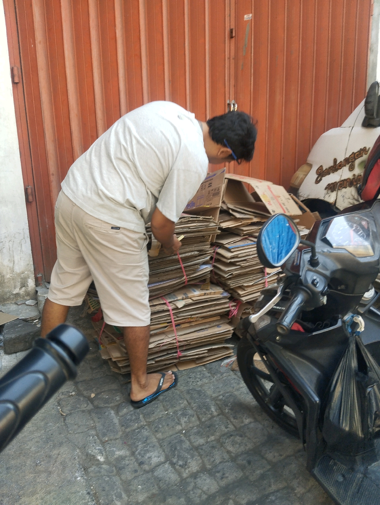 A man is bending over a stack of cardboard boxes in an outdoor setting. He is wearing a light-colored t-shirt and tan shorts. The cardboard boxes are stacked high and tied with pink string. Behind the man and the boxes is a ribbed, orange metal door. To the right of the boxes, a portion of a white vehicle with text on its side is visible, and a black motorcycle is parked in front of it. The ground is paved with interlocking stones. The scene appears to be in an urban environment, possibly a back alley or service area, characteristic of neighborhoods like Jodipan in Malang, Indonesia, known for its colorful houses and creative use of space, where such recycling activities might take place.