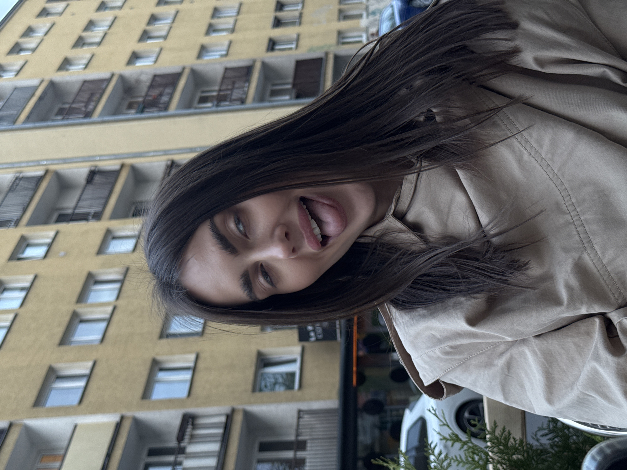 A young woman with long brown hair is sticking her tongue out at the camera. She is wearing a beige jacket or coat. Behind her is a tall yellow building with many windows, characteristic of Warsaw architecture. The scene likely takes place on a street in Warsaw, Poland.