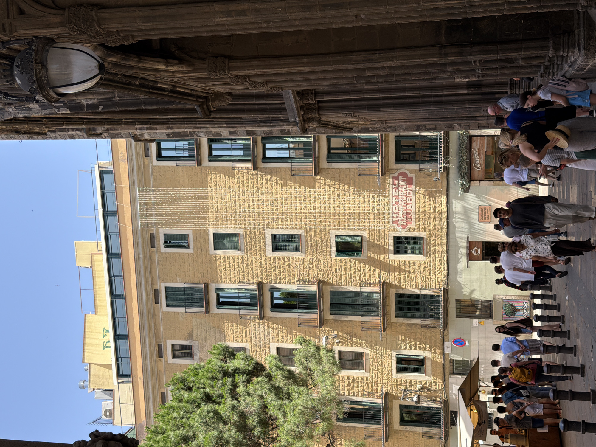 The image captures a lively street scene in Barcelona, Spain, bathed in bright sunlight, suggesting it is daytime. People are walking along a cobblestone street lined with buildings and shops. The architecture features a textured yellow facade on the main building, punctuated by numerous windows with dark green shutters, some open, revealing the interiors. Balconies with railings are also visible.
In the foreground, a large tree with dense green foliage partially obscures the view of the building. The street is lined with black bollards, and a market stall with a striped awning is set up, with several people gathered around it. Some individuals are looking at their phones, while others appear to be engaged in conversation or browsing.
Above, a stone archway with decorative carvings and a hanging lamp adds to the historic feel of the scene. The sky is a clear, bright blue, indicating good weather. A sign with red lettering on the yellow building reads "RESTAURANT" and below it, in smaller letters, "JARDÍ". Another sign on a nearby building appears to say "Giovanni". The overall atmosphere is one of a bustling urban environment on a sunny day.