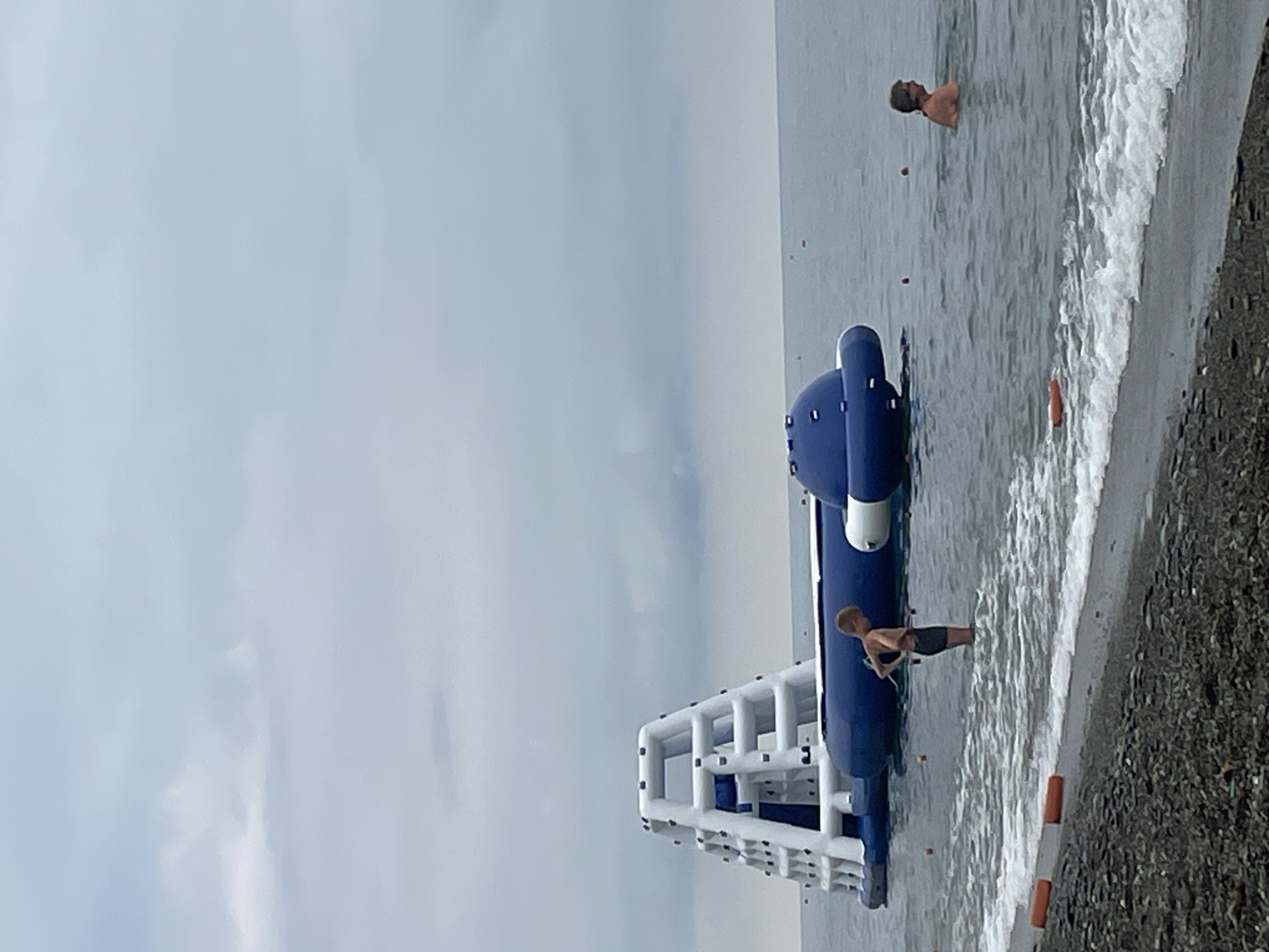 In Sochi, Russia, at an outdoor beach location, an inflatable water park attraction is situated in the sea. The attraction consists of a blue and white pyramidal climbing structure connected to a large blue inflatable with a domed section. Two individuals are visible in the water. A young boy, wearing dark swim shorts, stands in the shallow water near the shore, facing away from the camera and towards the water park. Further out in the sea, an adult, wearing only swimming trunks, is partially submerged in the water. The sea is calm with small waves lapping at the shore, which is covered in dark pebbles. The sky is overcast with muted, gray clouds. The overall atmosphere suggests a cloudy day at a recreational seaside area.