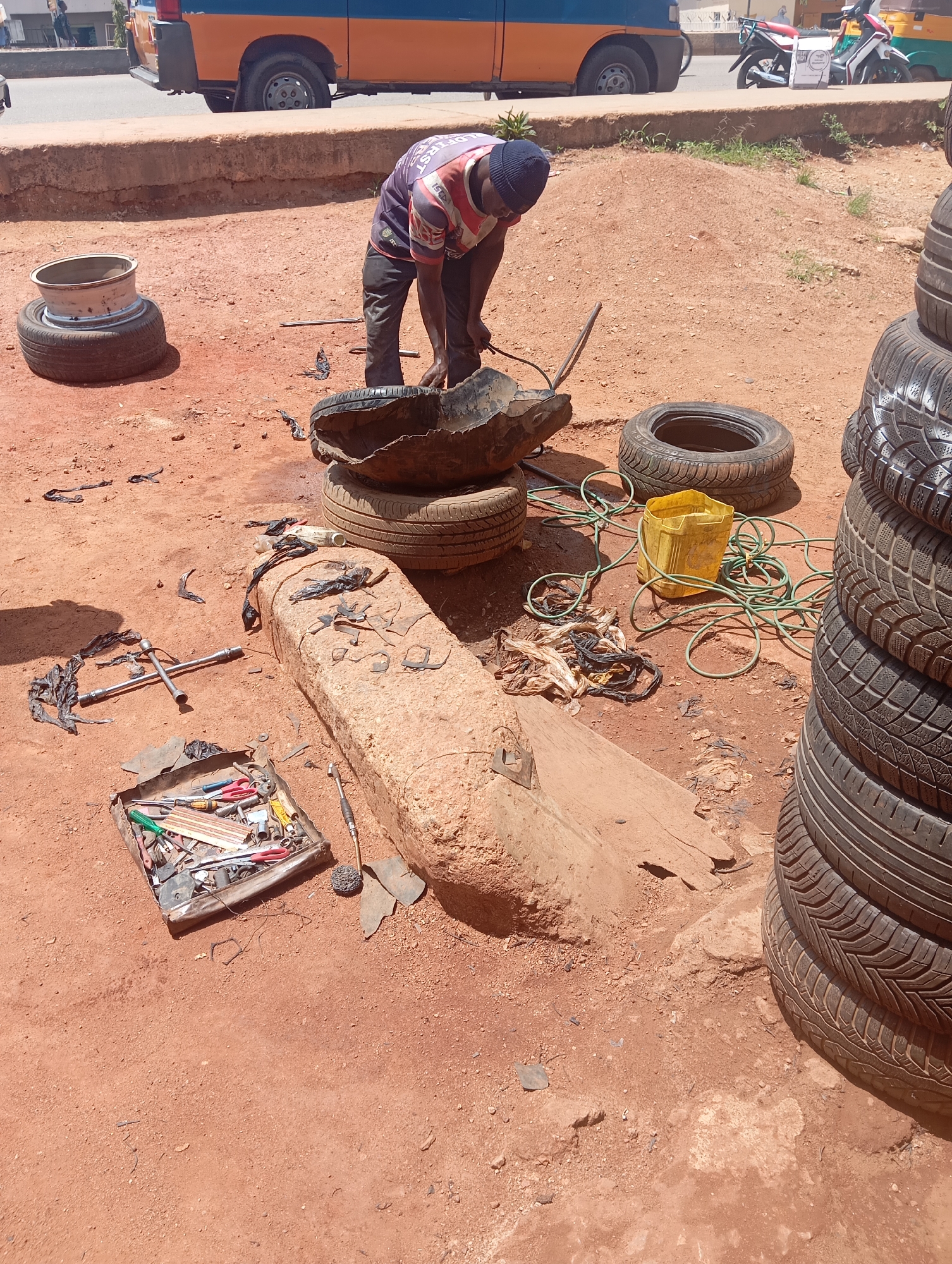 A man, wearing a blue beanie and a striped shirt with "ELDERST" visible on the back, is working on a tire outdoors. He is bent over, appearing to be either repairing or dismantling the tire. Several other tires are scattered around the scene, some stacked vertically and others lying on the ground. Tools are visible, including a wrench and a toolbox filled with various implements. A yellow bucket and a coiled green hose are also present.
The setting is dusty and appears to be a roadside workshop or informal repair area. The ground is covered in red dirt, and in the background, a truck with orange and blue panels is parked, along with a motorcycle. The lighting suggests it is daytime, with sunlight casting shadows. The overall impression is one of manual labor in a functional, no-frills environment. The presence of multiple tires and tools indicates a tire repair or recycling operation.