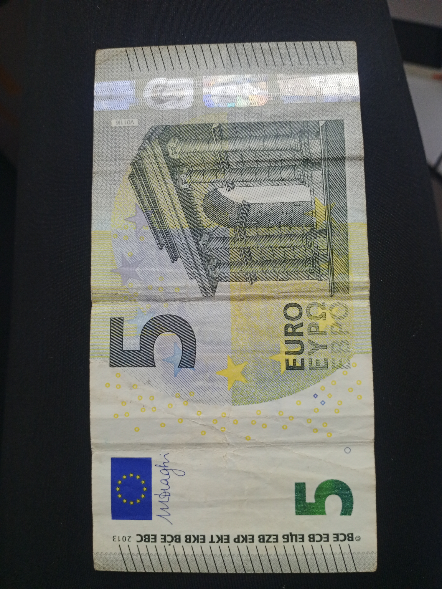 A vertical close-up shot displays a single 5 Euro banknote from the Europa series, likely resting on a dark, plain surface. The banknote appears slightly worn with visible creases, particularly across its center.
The design features a classical-style architectural element, resembling an aqueduct or bridge, predominantly in shades of grey and green. The denomination "5" is prominently displayed in the upper left and lower right corners, as well as in a larger font on the left side of the note's lower half. The word "EURO" is printed vertically on the right side, along with its Greek ("ΕΥΡΩ") and Cyrillic ("ЕВРО") equivalents. Yellow stars, part of the European Union flag motif, are visible on the left side of the note.
Visible security features include a holographic stripe at the top with a metallic sheen, containing the serial number "V01116" and other patterns. The signature of "M. Draghi" (Mario Draghi), former President of the European Central Bank, is present above the European flag symbol in the bottom left quadrant. Below this, the abbreviations for the European Central Bank in various languages are printed horizontally: "BCE ECB ЕЦБ EZB EKP EKT EKB BCЕ EBC 2013", indicating the series year. The background is uniformly dark and out of focus, highlighting the banknote as the sole subject.