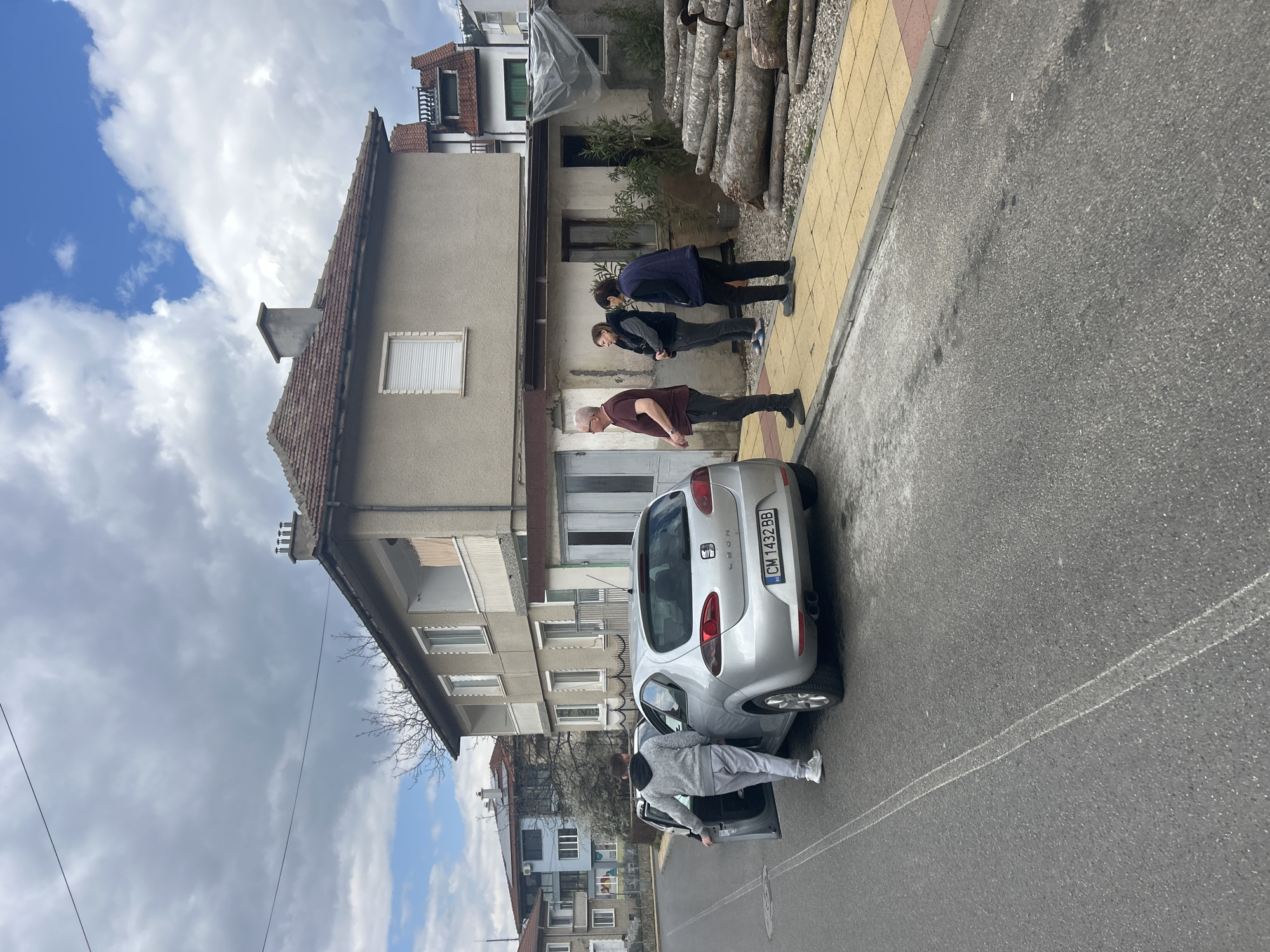 The scene is an outdoor urban setting in Zlatograd, Bulgaria. A silver Seat Leon car, with license plate "CM 1432 BB", is parked on the right side of an asphalt road near a sidewalk. A person in a gray sweatshirt and light pants is seen exiting the car. Three people stand on the sidewalk: one in a maroon shirt and black pants, and two others in dark clothing. A residential building is adjacent to the sidewalk, with a pile of logs stacked near the building. The sky above is partially cloudy with patches of blue.