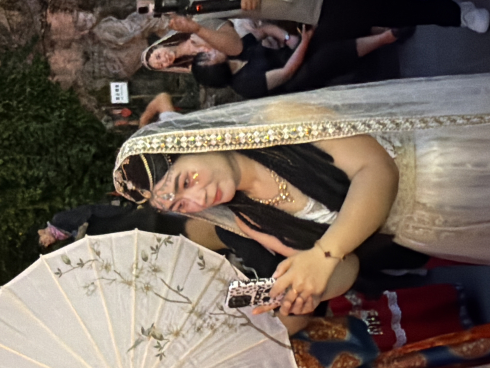 In Yuzhong District, China, a woman is seen holding a mobile phone in her left hand while posing for a picture. She is wearing a white veil with gold trim, a beaded necklace, and a white dress. The background is a rocky cliff face with some greenery. Other people are visible in the background, including a woman in a similar veil and attire, and several other individuals in casual clothing. A white umbrella with a floral design is held by someone to the left of the main subject. The lighting suggests it is evening or indoors with artificial lighting.