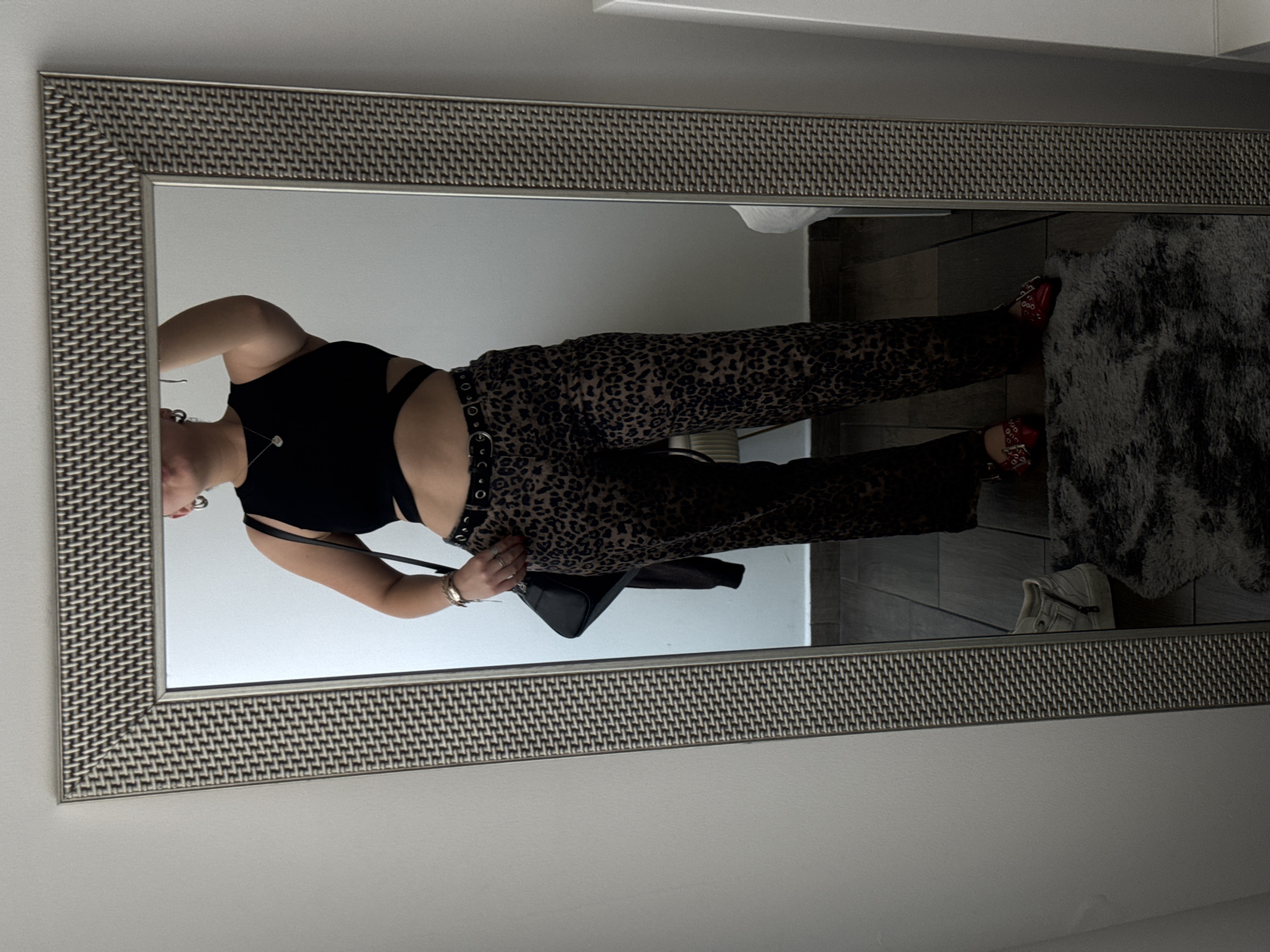 A person is captured in a mirror selfie, standing indoors and posing. The image shows the person from approximately the mid-chest down to their feet, reflected in a large, rectangular mirror. The mirror has a distinctive silver-toned frame with a woven or textured pattern.
The person is wearing a black sleeveless crop top that reveals their midriff, paired with high-waisted leopard print pants. A black belt with multiple silver rings is cinched around their waist. On their feet are red shoes with multiple straps and silver buckle detailing. A black handbag with a structured shape is held in one hand, near their hip. Visible accessories include a simple necklace with a small white pendant, earrings, a watch on one wrist, and rings on their fingers.
In the reflection within the mirror, besides the person, parts of the room are visible. To the right of the person's feet, a grey furry rug is seen on what appears to be a tiled floor. Further in the background reflection, a white sneaker or casual shoe is visible near the floor. The lighting suggests an indoor setting, possibly a room like a bedroom or dressing area. There is no visible text in the image.