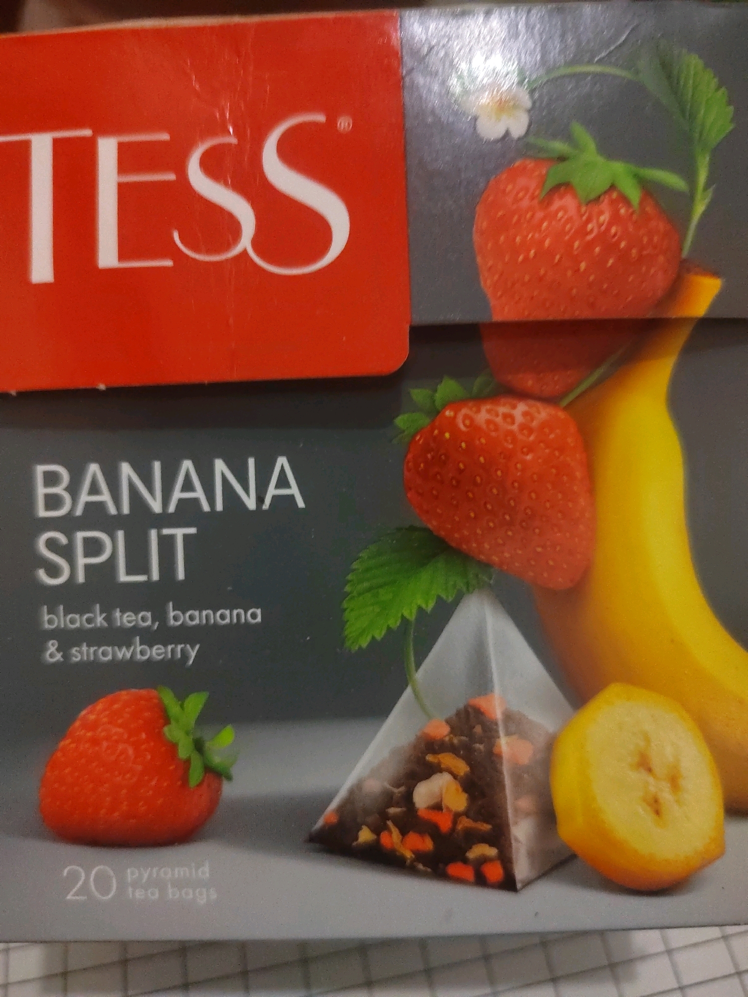 This is a product shot of TESS Banana Split black tea. The packaging features the TESS logo in white text on a red background. Below this, "BANANA SPLIT" is written in large white letters, followed by "black tea, banana & strawberry" in smaller white text. The image displays several visual elements: two strawberries with green leaves and a small white flower, a portion of a banana, and a pyramid-shaped tea bag filled with tea leaves and fruit pieces. A slice of banana is also visible. The number "20 pyramid tea bags" is printed in white text at the bottom left. The background is a dark gray gradient. There are no people or any indication of a specific location, including in the city of Bolshaya Elhovka, Russia.