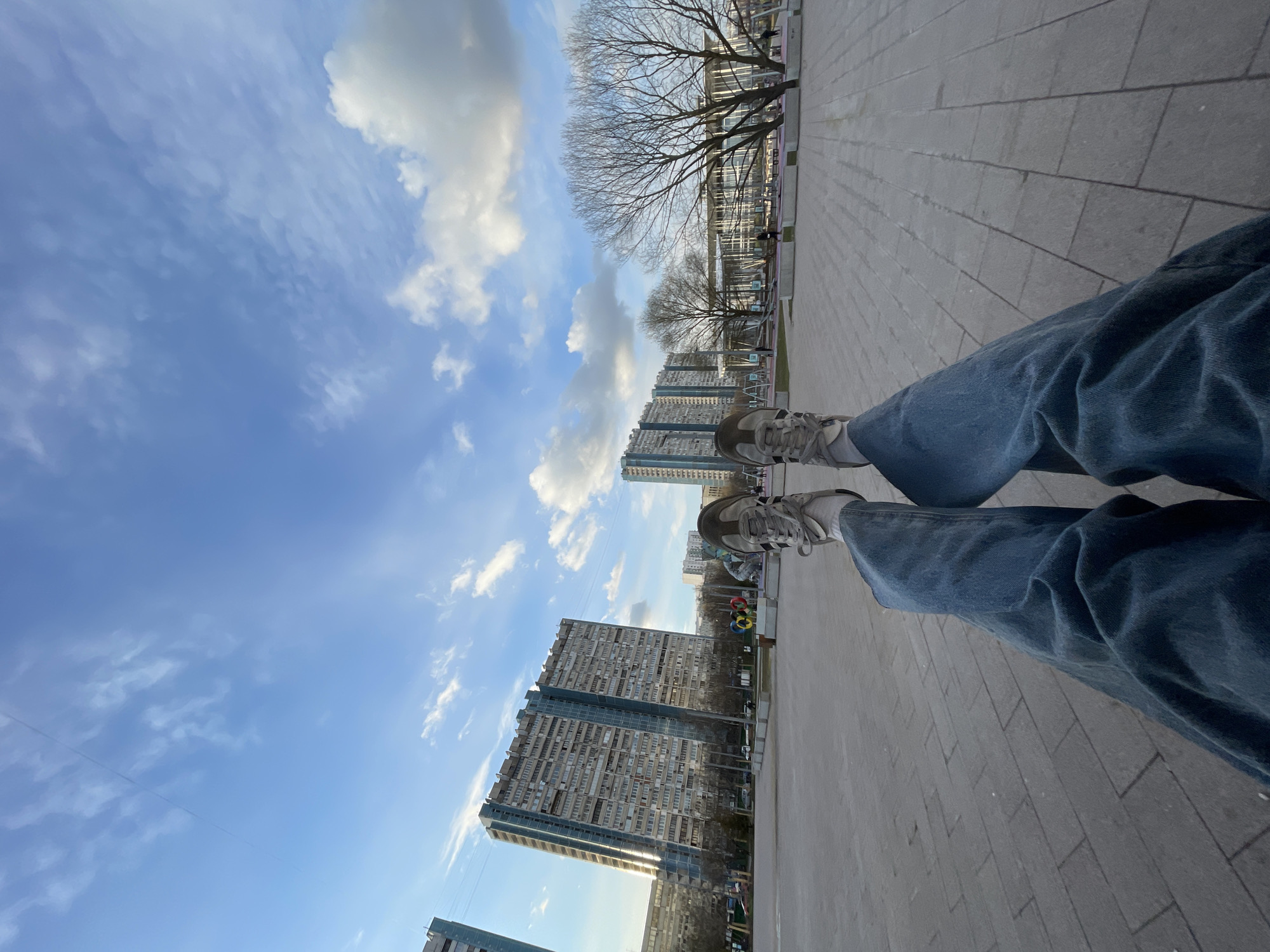 The image depicts a first-person perspective of someone sitting with their legs outstretched, wearing blue jeans and sneakers, in an urban environment. The ground is paved with rectangular gray stones. Several tall, Soviet-era apartment buildings are visible in the background, suggesting a location in a residential district. The sky is blue with scattered white clouds. Bare trees line the walkway near what appears to be a playground or recreational area with colorful installations. The overall setting points to a public space, possibly a park or plaza in Moscow, Russia, recognizable due to the architectural style of the buildings and urban planning. There is no interaction happening in the image.