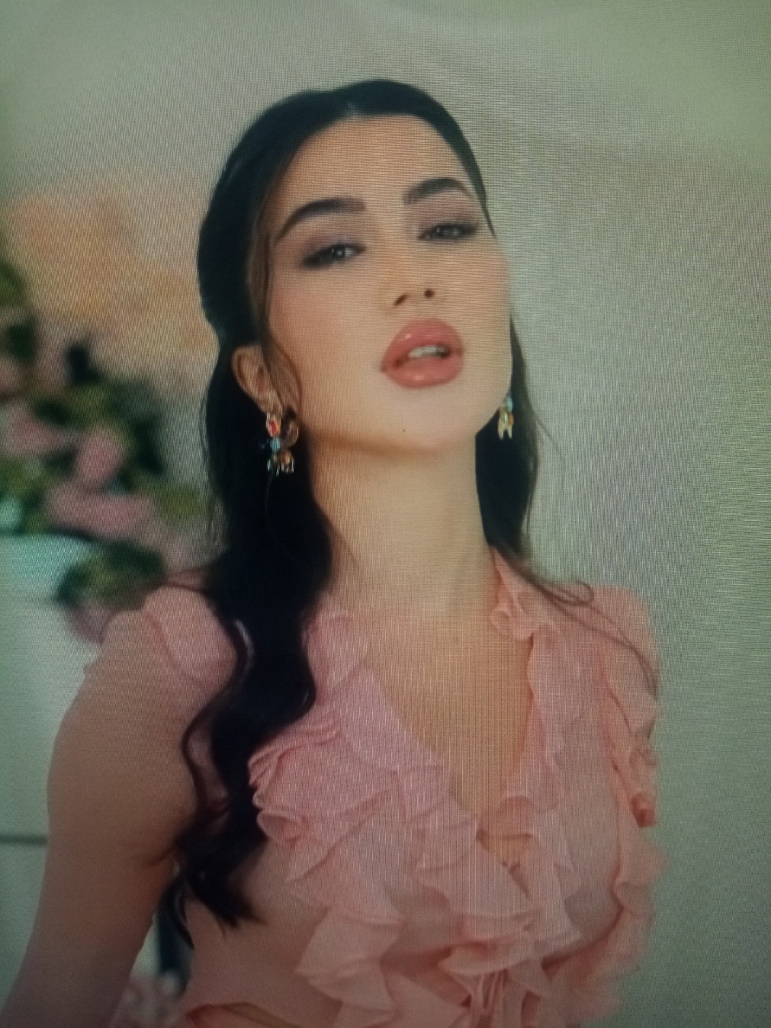 A woman with dark hair and full lips, wearing a pink ruffled shirt and colorful earrings, poses for a photo in what appears to be a dimly lit indoor setting. The background is blurred, with hints of floral arrangements on the left side. The image is of low resolution and appears to be a photograph of a screen, showing scan lines across the surface. The subject's facial expression is suggestive and alluring. No elements within the image suggest a connection to the city of Chirakchi, Uzbekistan.