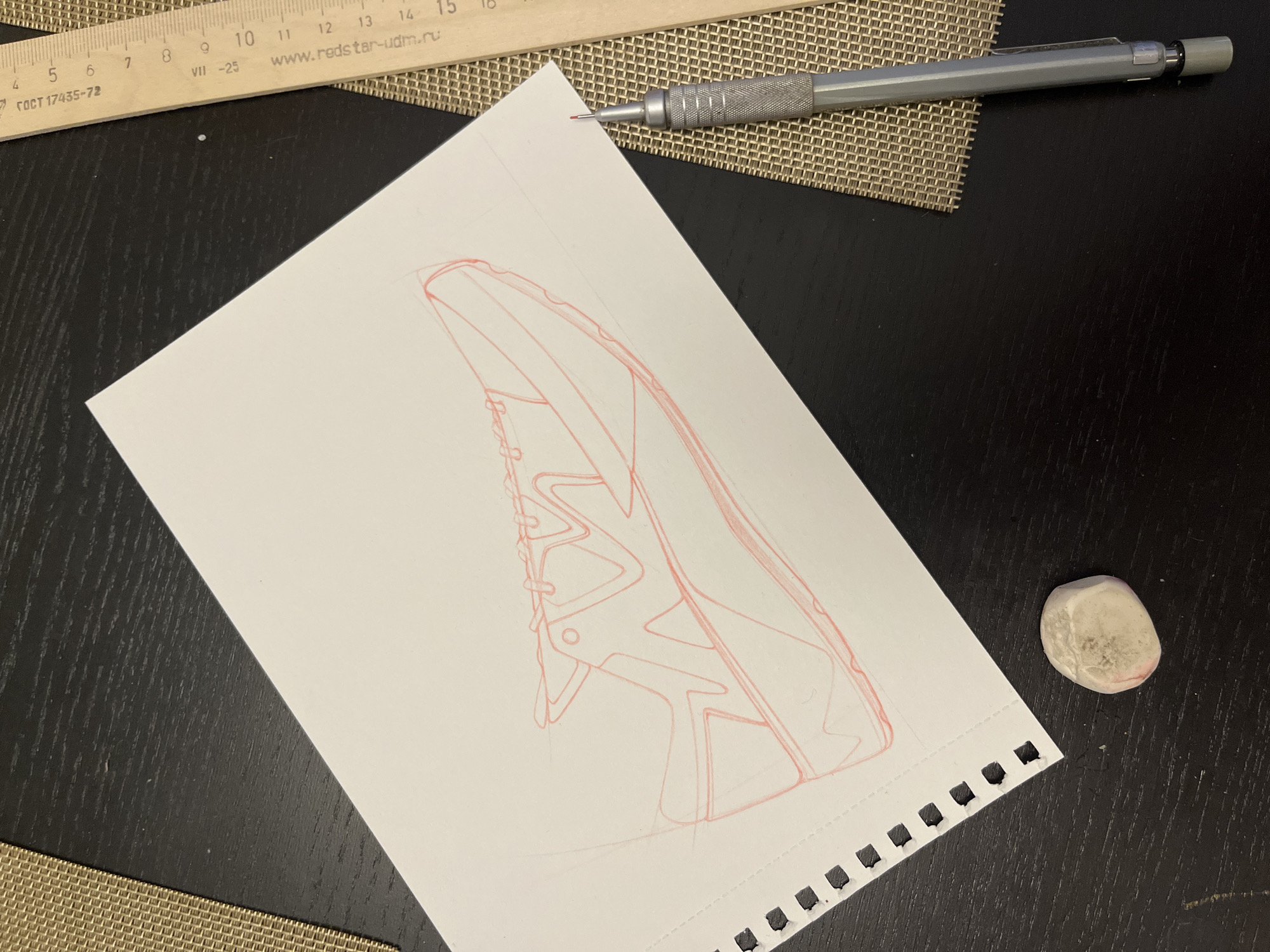 The image depicts a drawing of a shoe on a piece of white, perforated paper. The drawing is done in red ink and appears to be a preliminary sketch. A grey mechanical pencil rests near the top of the paper, partially on a woven placemat. A wooden ruler is positioned at the top edge of the image, with markings in centimeters. An eraser sits to the right of the paper. The background is a dark, possibly black, textured surface.