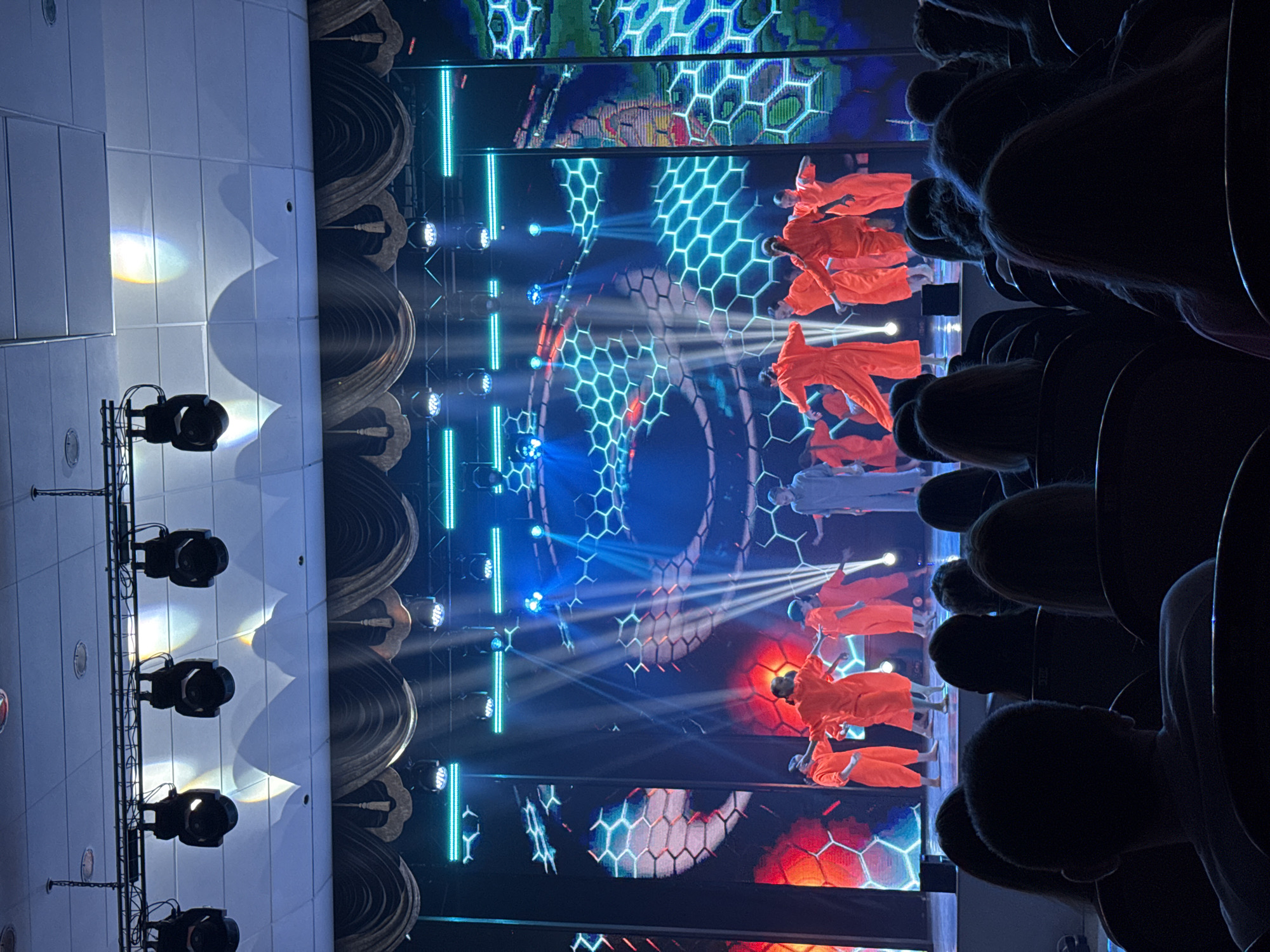 Here is a description of the image:
The image shows a performance on a stage, likely indoors. The view is from the audience. Several performers are visible, wearing orange robes and appearing to float or fly in the air, aided by wires or harnesses. A single performer appears to be wearing a white robe. The stage backdrop features a large screen displaying a hexagonal pattern in blue and red. Bright stage lights illuminate the performers and the backdrop. The audience is visible in the foreground. There are lights hanging from a bar on the left side of the stage. Without additional data, it's impossible to confirm whether the scene is specifically in Krasnoyarsk, Russia.