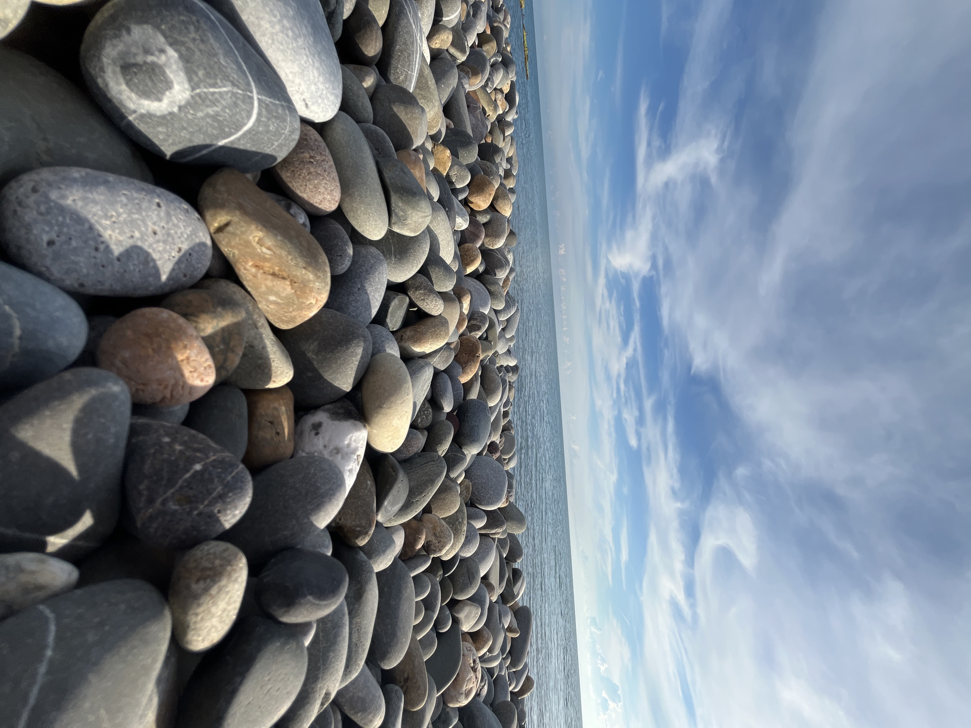 This is a low-angle, wide shot of a pebble beach, likely in Sochi, Russia, under a bright, partly cloudy sky. The foreground is dominated by a dense arrangement of smooth, rounded pebbles in various shades of gray, brown, and white. Some pebbles are larger and more detailed, showing striations and patterns, while others are smaller and more uniform. The pebbles extend towards the middle ground where they meet the calm, blue sea. The horizon line is distinct, with the ocean meeting the sky. The sky itself is a vibrant blue, streaked with thin, wispy white clouds that are illuminated by sunlight, creating a dynamic visual effect. The overall impression is one of a serene, natural coastal landscape. There are no people or animals visible in the image.