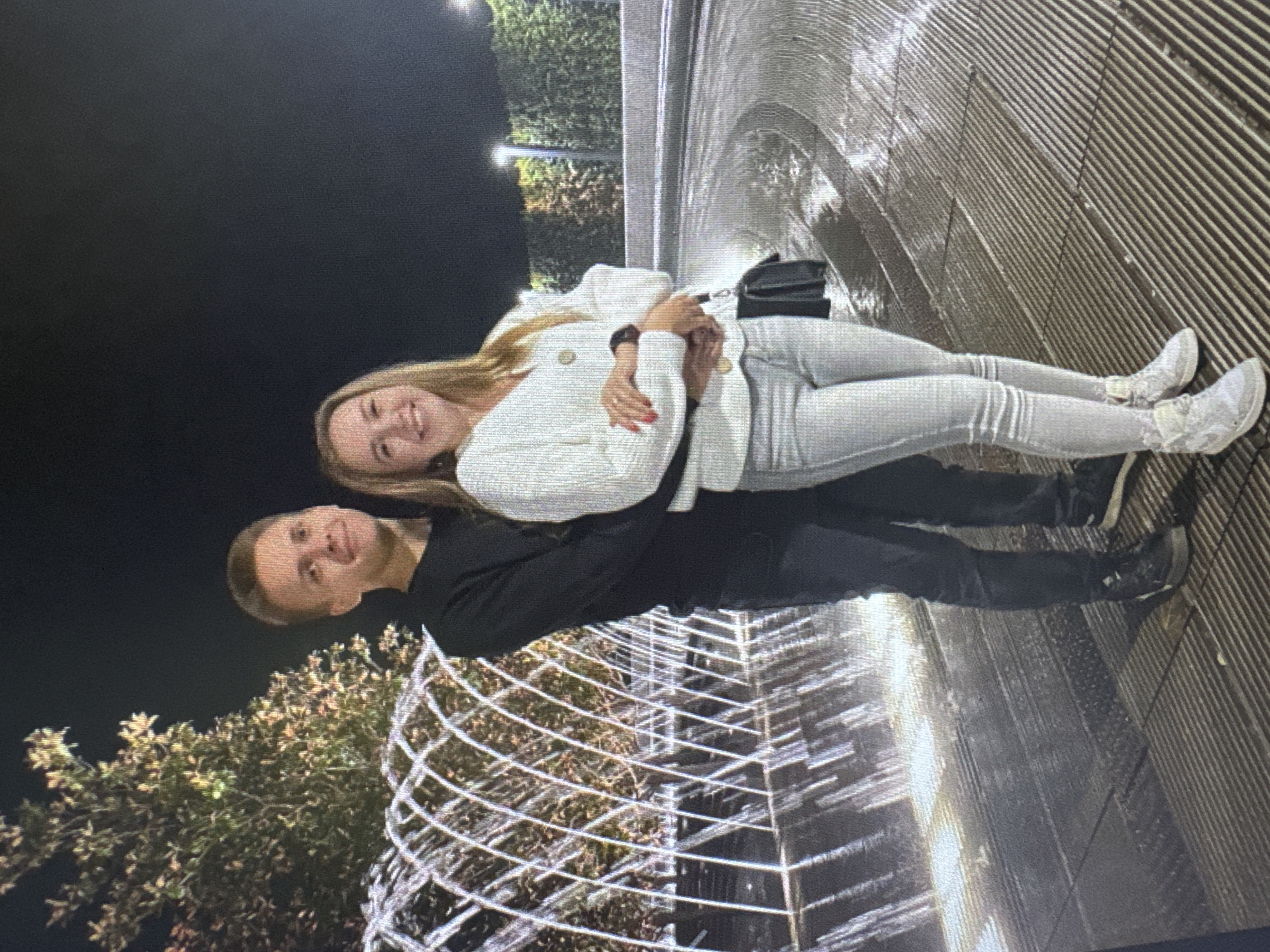 A young adult couple stands embracing in front of a fountain at night. The male, on the left, wears a dark long-sleeved shirt and dark pants. The female, on the right, wears a white V-neck sweater with gold buttons and light-colored pants. She has blonde hair and is smiling. Both are wearing sneakers. The fountain features curved structures with water spraying from them, creating reflections on the wet ground. The background is dark, with some illuminated trees and structures suggesting an urban park or plaza setting.