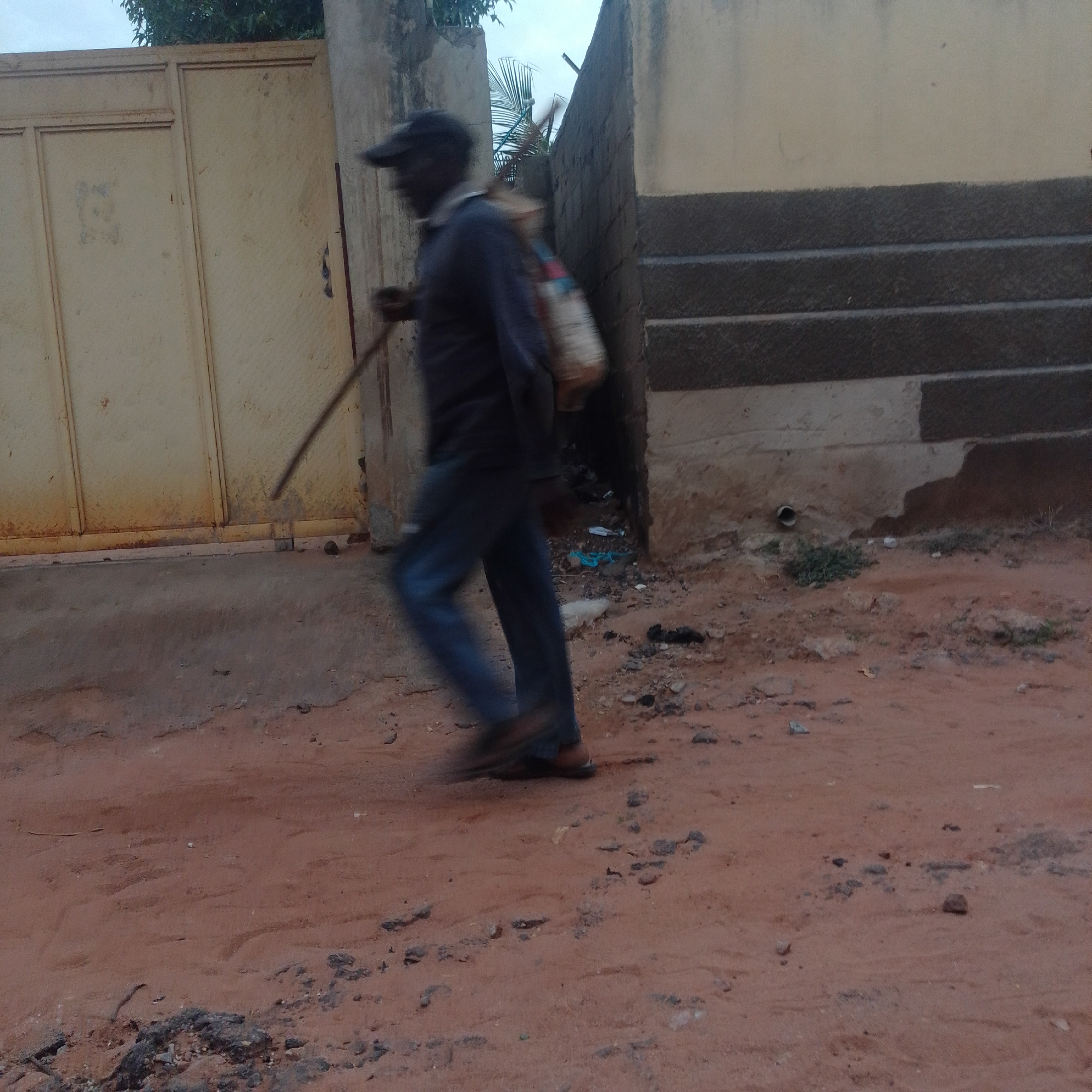An unidentified person, wearing a dark baseball cap, a dark long-sleeved shirt, and blue jeans, walks along a dirt path carrying a light-colored bag slung over their shoulder and holding a long, thin object, possibly a broom or stick, in their right hand. The person is in motion, as indicated by the motion blur. The background features a pale yellow metal gate on the left and a textured concrete wall with dark gray horizontal bands on the right. The ground is a reddish-brown dirt path with scattered pebbles and debris. The sky above is overcast. The scene suggests an outdoor urban or semi-urban environment.