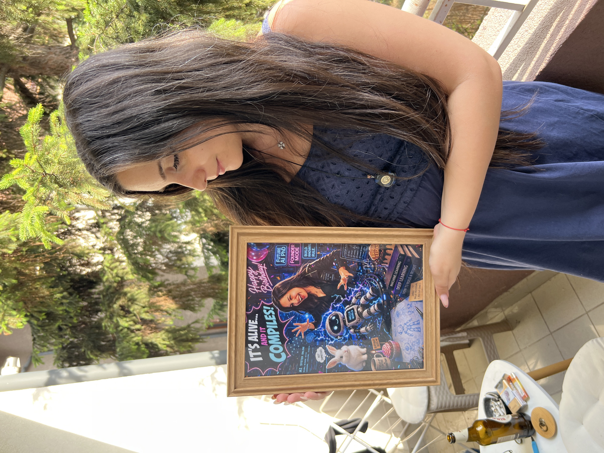 In Sofia, Bulgaria, a young woman with long, dark brown hair and a blue dress stands on a balcony, holding a framed picture. The picture depicts a woman with dark hair and a black shirt, smiling and gesturing excitedly, with a robotic figure and a white rabbit next to her. Text on the picture reads "IT'S ALIVE... AND IT COMPILES! Happy Birthday!" and mentions "FUTURE AI PHD" and "FOUNDER MODE." The background of the image shows lush green trees and a railing, suggesting an outdoor setting. On a small white table to the left, a bottle of beer, a pack of cigarettes, and other items are visible. The lighting and shadows indicate it is daytime.