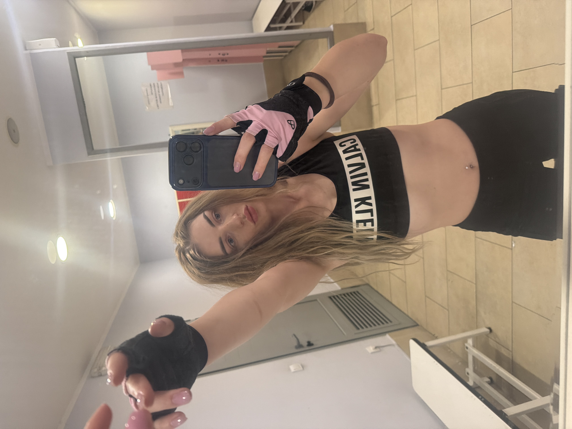 In an indoor setting, a young woman with blonde hair takes a selfie in a mirror. She is wearing a black sports bra with white lettering, black athletic shorts, and fingerless black and pink workout gloves. Her left arm is raised, and she is holding a smartphone in her right hand. The background suggests a locker room or gym changing area, with tiled flooring, a grey door with a vent, a white bench, and a large mirror reflecting the scene.