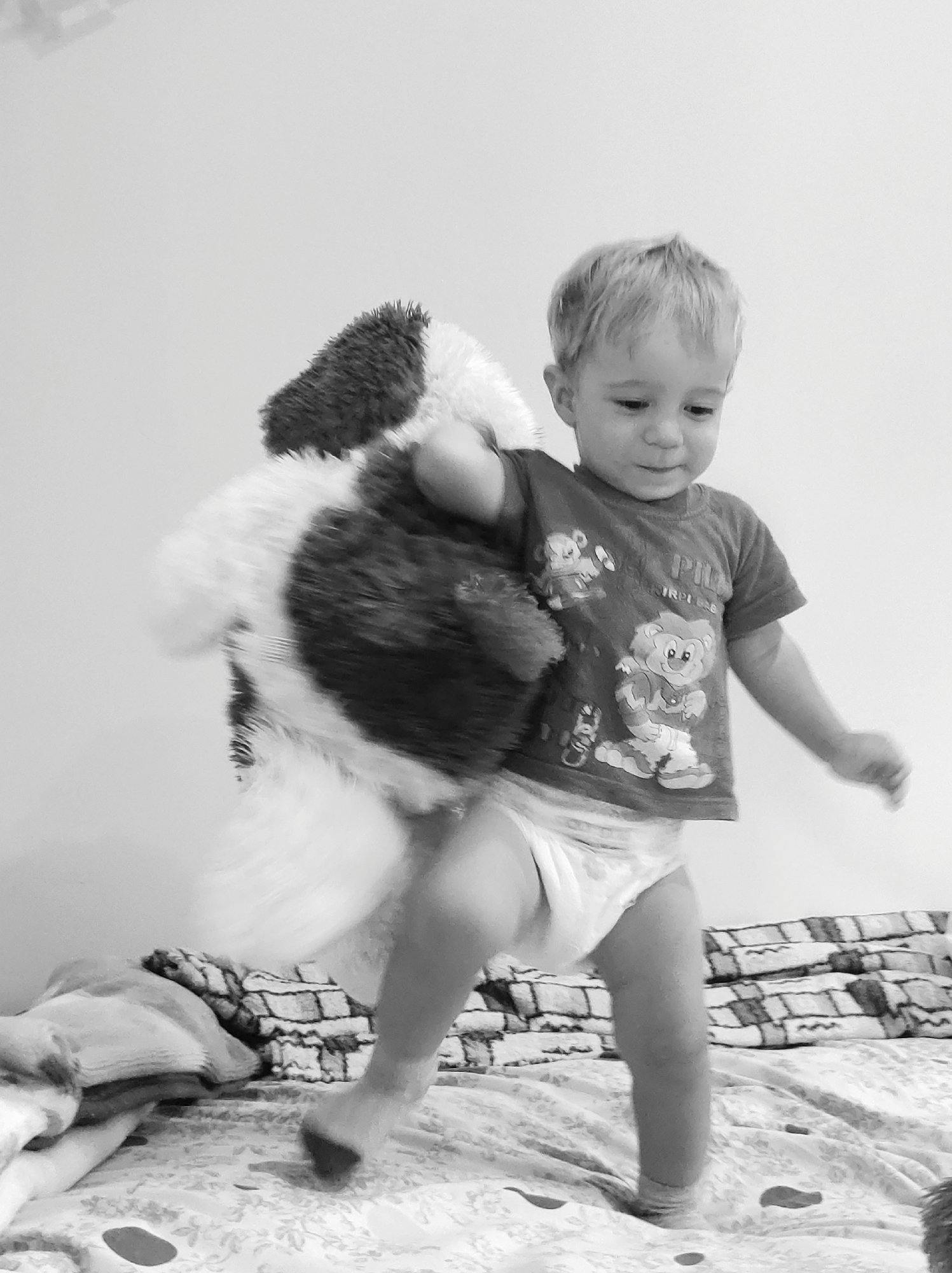 A monochrome, medium-shot photograph captures a toddler actively engaged in play. The child, facing towards the right and slightly downwards, is holding a large, plush stuffed animal, possibly a dog, that is predominantly white with black patches. The toddler wears a t-shirt adorned with cartoon bear graphics and a diaper. Their left leg is bent and extended forward, suggesting movement. The setting appears to be a bedroom, with a bed visible in the foreground covered with patterned bedding and a stack of blankets or pillows to the left. The background consists of a plain, light-colored wall.