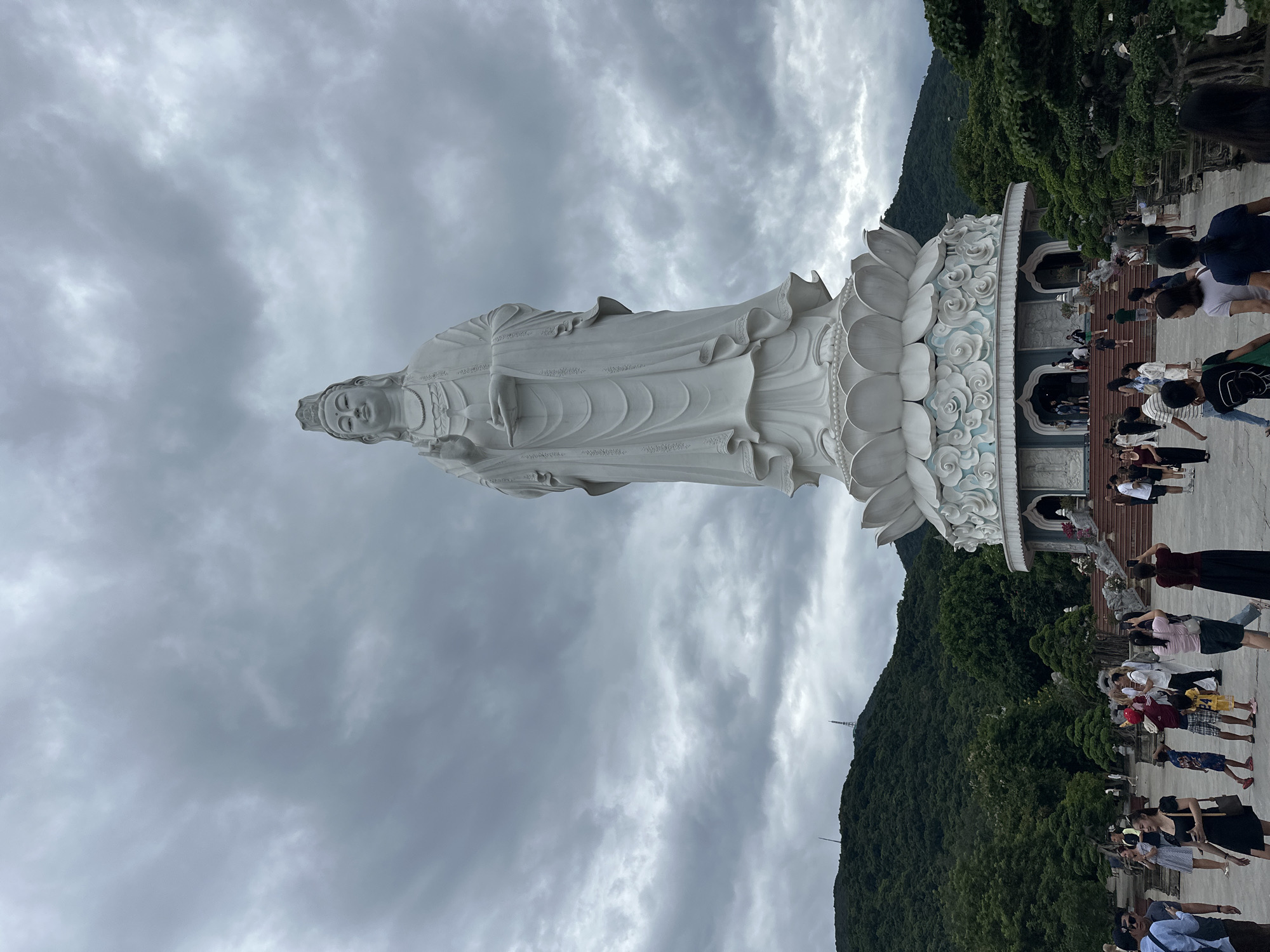 The image shows a large, white statue of a Buddhist figure, likely Guan Yin, standing on a lotus flower pedestal. The statue is the central subject, with its right hand raised in a blessing gesture and its left hand holding a small object. The statue is located in a courtyard area where a crowd of people are gathered. Some individuals are taking pictures, while others are walking around.
Behind the statue, a lush green, tree-covered mountain rises. The sky above is overcast with dramatic grey clouds. In the distance, on the mountain's crest, two telecommunication towers are visible. The courtyard itself is paved, with decorative landscaping, including trees in planters, lining the sides. Architectural elements, possibly part of a temple or religious complex, are visible at the base of the statue's pedestal, featuring arched entrances and relief carvings.
The location is the Lady Buddha statue (Chùa Linh Ứng) on Son Tra Peninsula in Da Nang, Vietnam. The scene captures a popular tourist and pilgrimage site on an overcast day.