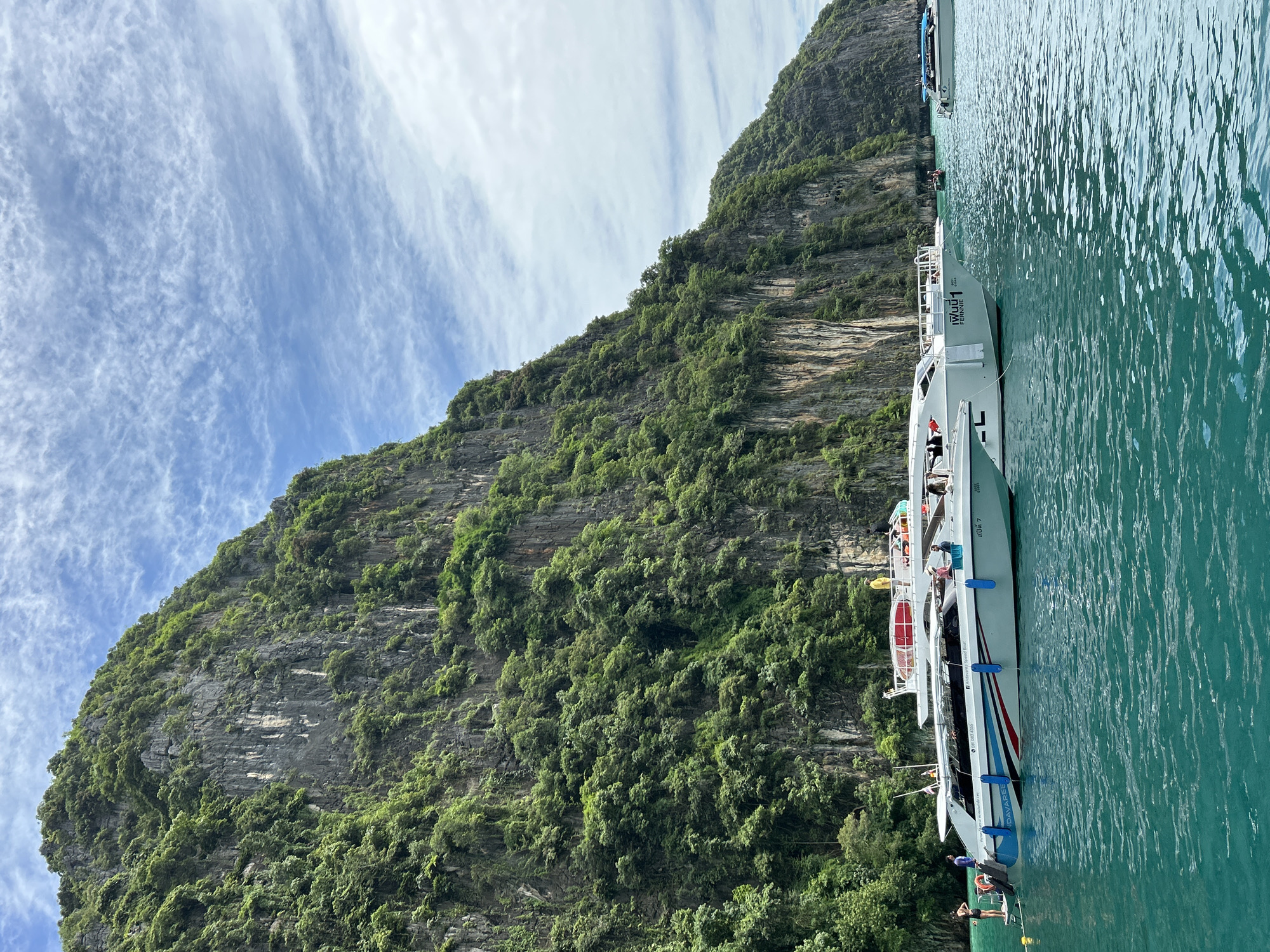 The image depicts a scenic view of limestone karst cliffs covered in lush green vegetation, rising dramatically from the turquoise waters of a bay. Several speedboats are anchored near the shore. In the foreground, a white speedboat with blue and red accents is prominently featured, with people visible on its deck and boarding via a yellow floating buoy. Another white speedboat is docked behind it, and a smaller boat can be seen further in the distance. The sky is partly cloudy, with wispy white clouds against a blue backdrop. The overall scene suggests a popular tourist destination in Thailand, likely a coastal area known for its natural beauty and maritime activities.