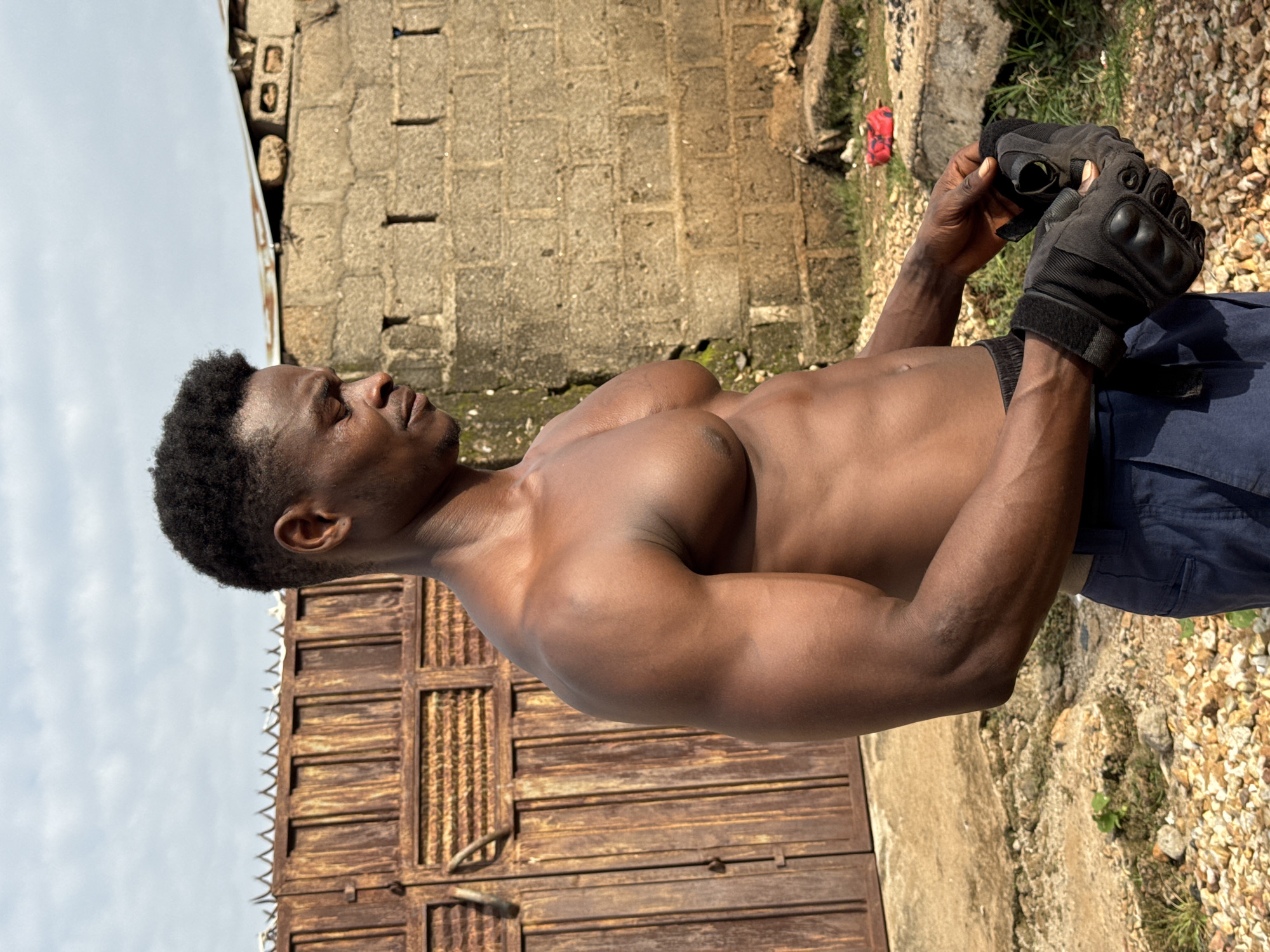 A shirtless Black man with dark skin and short, dark, curly hair is standing outdoors in what appears to be an urban or semi-urban environment. He is looking down and to his right, with his eyes closed and his head slightly bowed. He is wearing dark blue pants and black fingerless gloves. He is holding the gloves in his hands, as if he is putting them on or taking them off. The background consists of a weathered, rusty metal gate on the left, a rough brick wall on the right, and a cloudy sky above. The ground is covered with gravel and scattered debris. The overall impression is one of an individual engaged in some form of physical activity or preparation. The location is identified as the city of Akwanga, Nigeria.