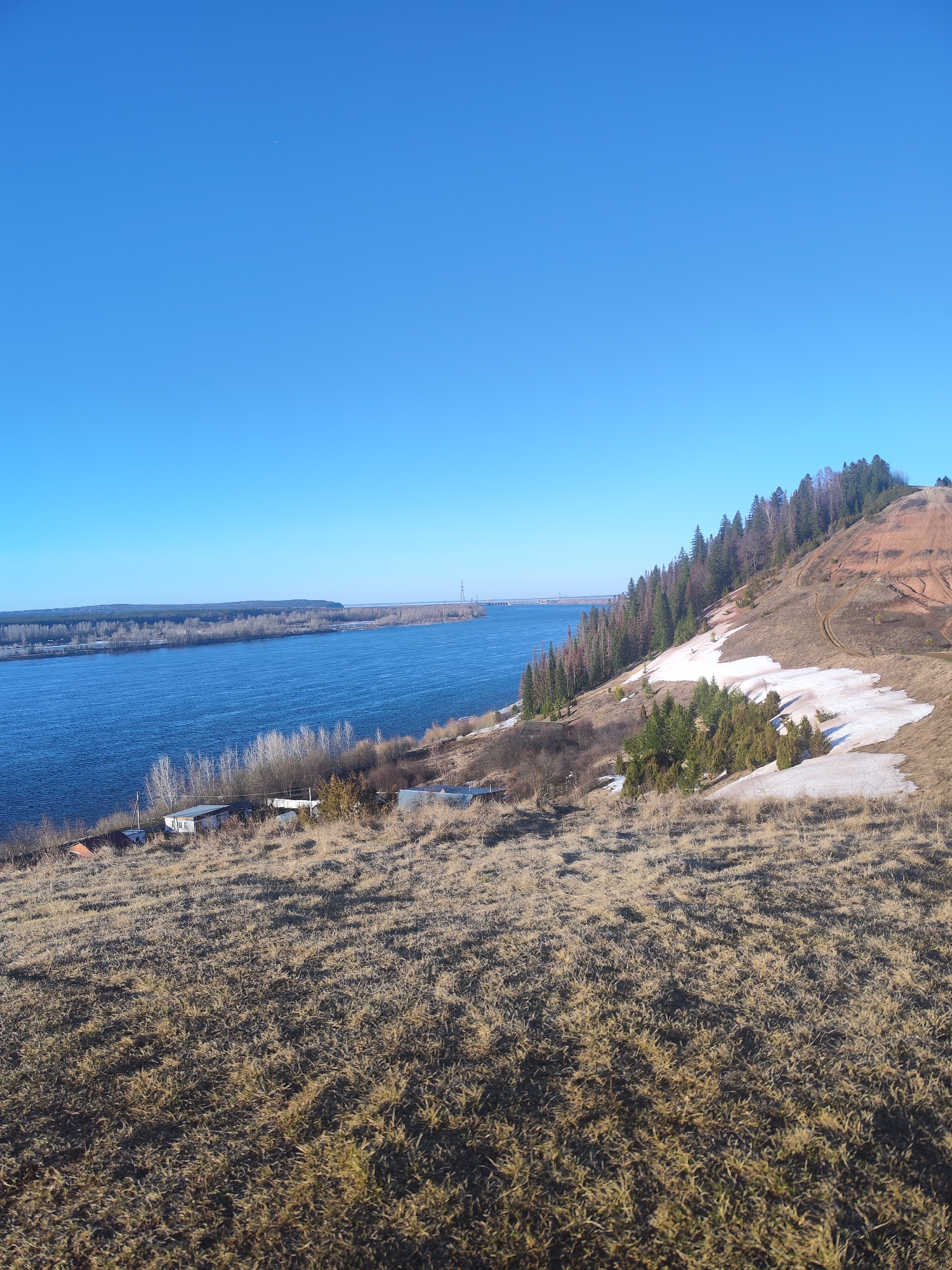 The image depicts a wide river, likely the Volga, with a forested island in the distance and a bridge spanning it. The scene is viewed from a high vantage point on a hill, covered in dry grass with patches of melting snow. Coniferous trees line the hillside, transitioning to a red-toned, barren upper slope. A few buildings are visible near the riverbank, nestled among sparse vegetation. The sky is clear blue. The location is likely in the vicinity of Noviy, Russia, considering the river's characteristics and the landscape.