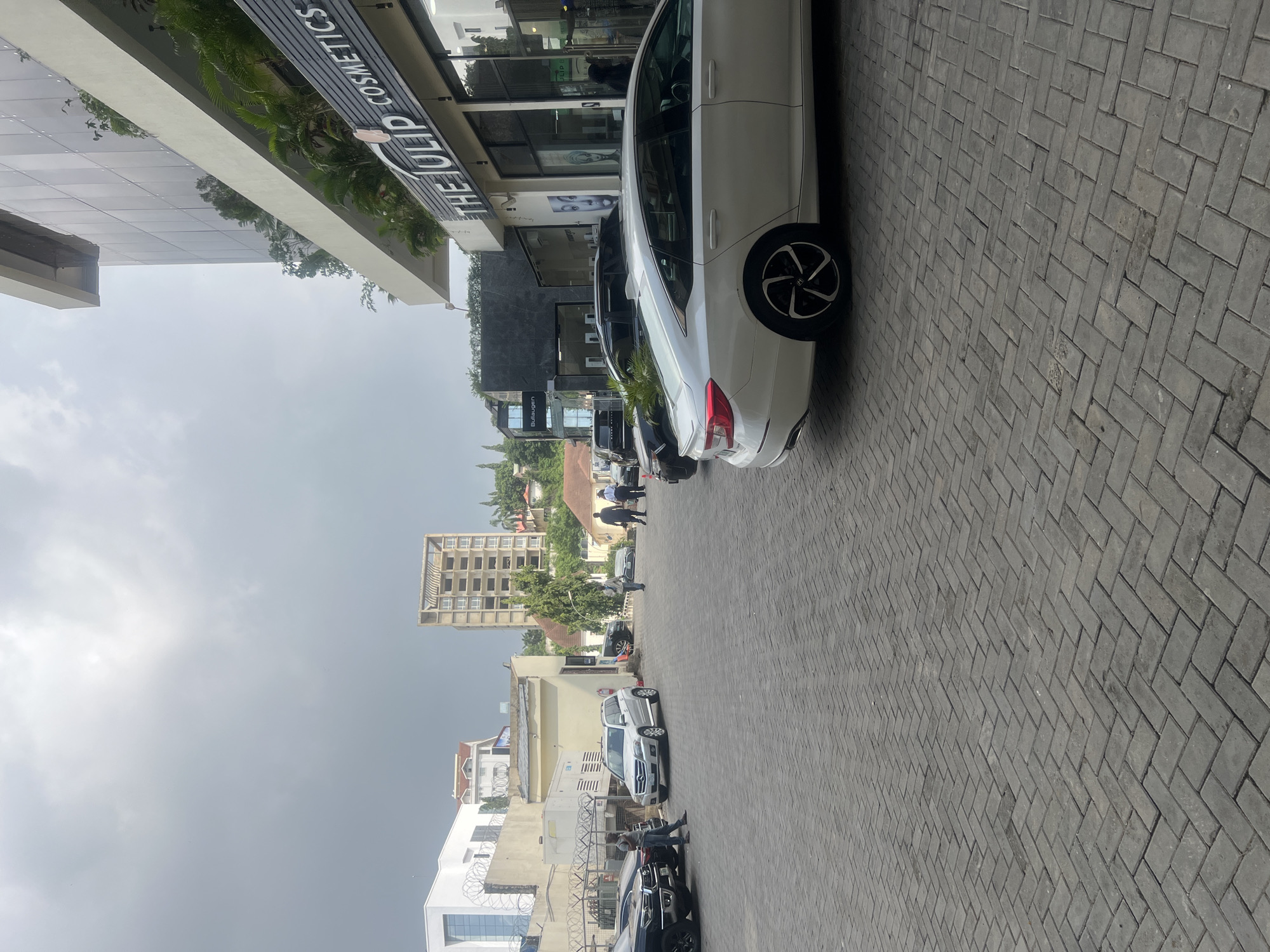 The image depicts an exterior view of a modern commercial building, likely in Abuja, Nigeria. The foreground is dominated by a paved driveway or parking area made of interlocking gray bricks laid in a herringbone pattern. Several vehicles are present, including a white sedan in the mid-right of the frame, a silver SUV, and the front of a black SUV. A few individuals are visible in the scene: two men in dark suits are standing near a black SUV on the right, and a man in a light-colored shirt and jeans is standing near the silver SUV on the left. In the background, various buildings are visible, including a multi-story building with a glass facade that houses "THE TULIP COSMETICS" store, indicated by signage. Other buildings include a white structure with security fencing and a taller, partially constructed building. The sky is overcast, suggesting a cloudy day. The overall scene conveys a sense of urban commercial activity.