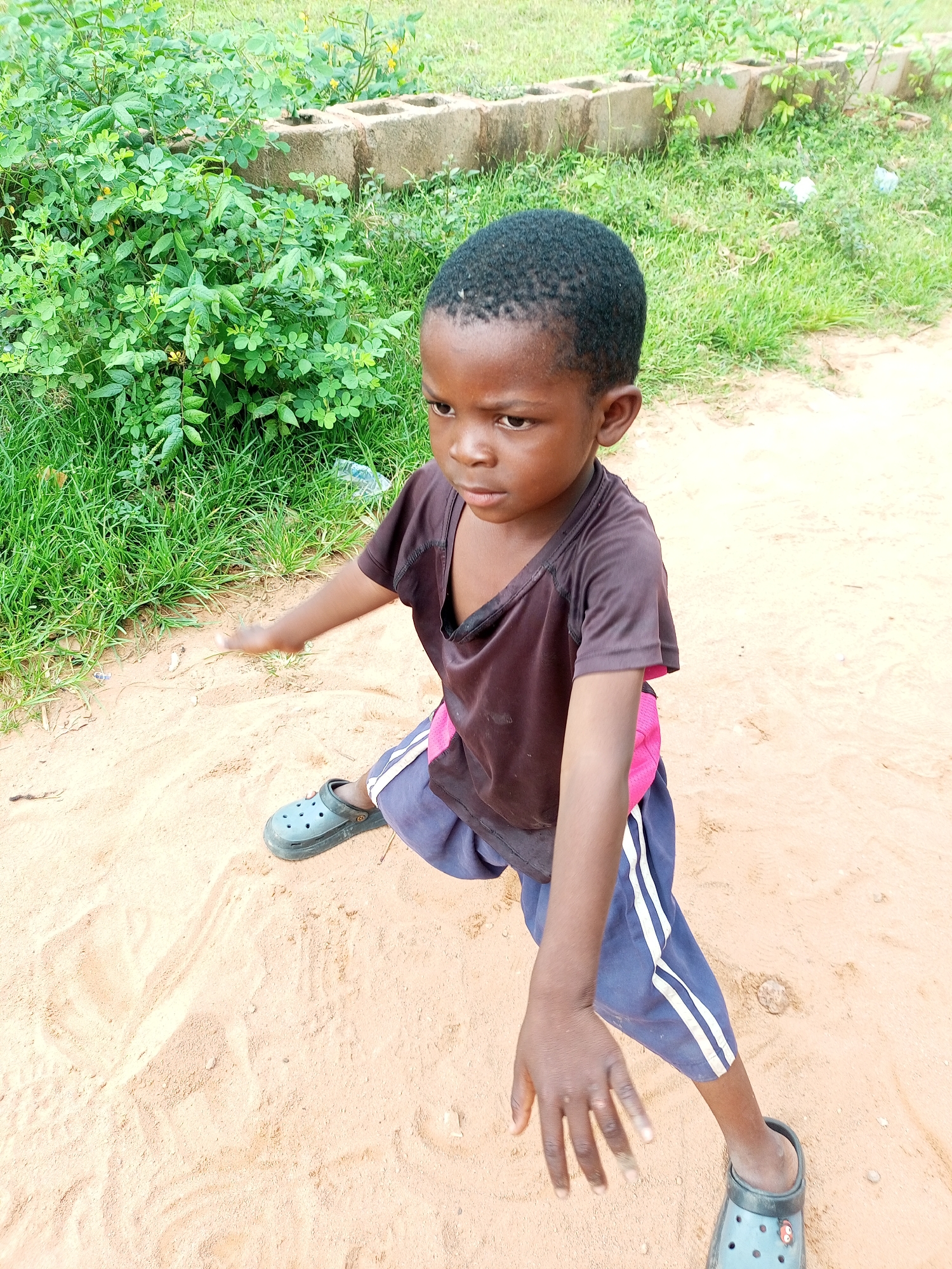A young boy is seen from a high angle, sitting on a sandy surface with a lush green bush and some concrete blocks in the background. The boy is wearing a dark t-shirt and blue shorts with white stripes. He has dark skin and short black hair. His left leg is bent, and his right leg is extended. His arms are outstretched, with his left hand reaching forward. He is wearing a dark-colored shoe on his left foot. The environment appears to be an outdoor setting, possibly a rural area given the vegetation and sandy ground. The lighting suggests it is daytime.