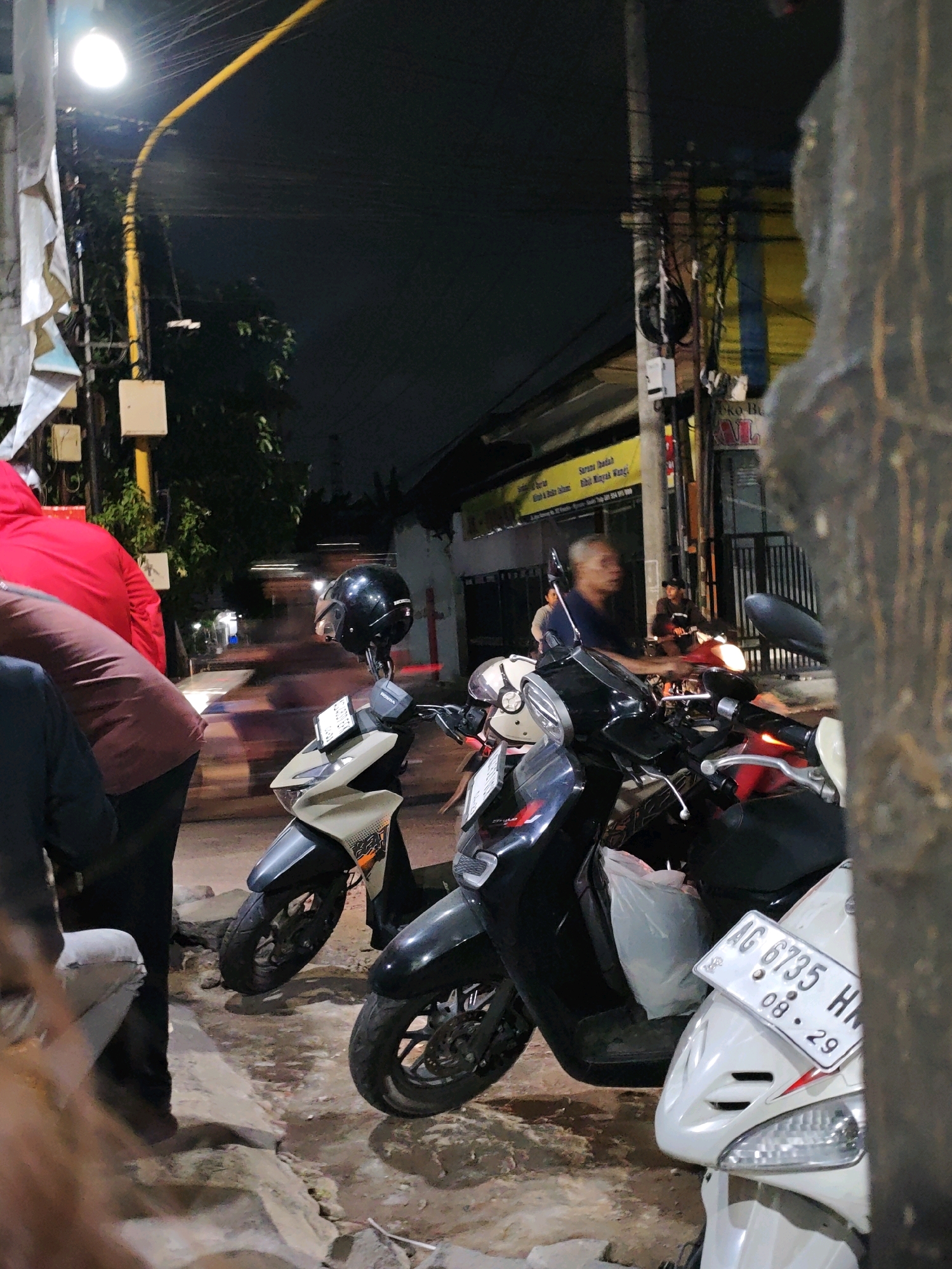 The image captures a street scene at night in Ngasem, Indonesia, focusing on parked motorcycles and a few individuals. In the foreground, a light-colored motorcycle with a license plate reading "AG 6735 HN 08-29" is partially obscured by a tree trunk on the right. To its left, a black scooter is parked, with a white helmet resting on its handlebars. Further left, a person wearing a red jacket and brown pants is standing. In the background, a man with a blurred face is riding a motorcycle, his face lit by the vehicle's headlight. Another person is visible on a motorcycle behind him. The environment appears to be a street with buildings and overhead wires, illuminated by streetlights and the headlights of vehicles. The ground is rough and uneven, with rocks and debris. The overall impression is a snapshot of nocturnal street life in a moderately urban setting.