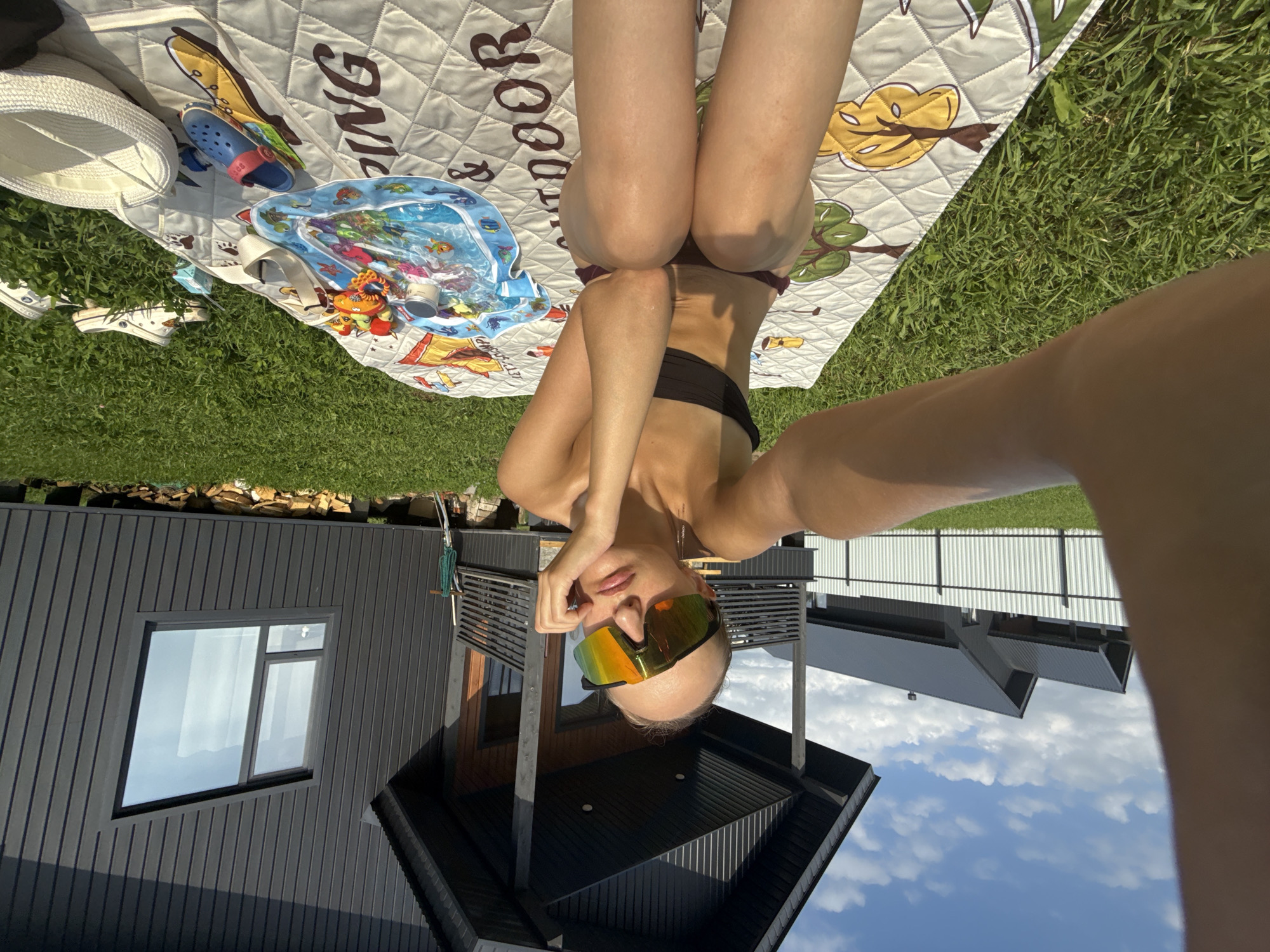 The image depicts a person lying on a patterned blanket on a grassy lawn. The person is wearing a bikini and large, reflective sunglasses. The scene is set outdoors on a sunny day, indicated by the bright light and shadows. In the background, there is a modern dark-colored building with a large window and a sloped roof. The sky is blue with some scattered clouds. There are also some children's items on the blanket, including a small inflatable pool and a pair of blue and red sandals, suggesting a recreational or family outing. The overall mood appears relaxed and summery. The text "KING" is visible on the blanket. The location context provided is Podshivalovo, Russia.