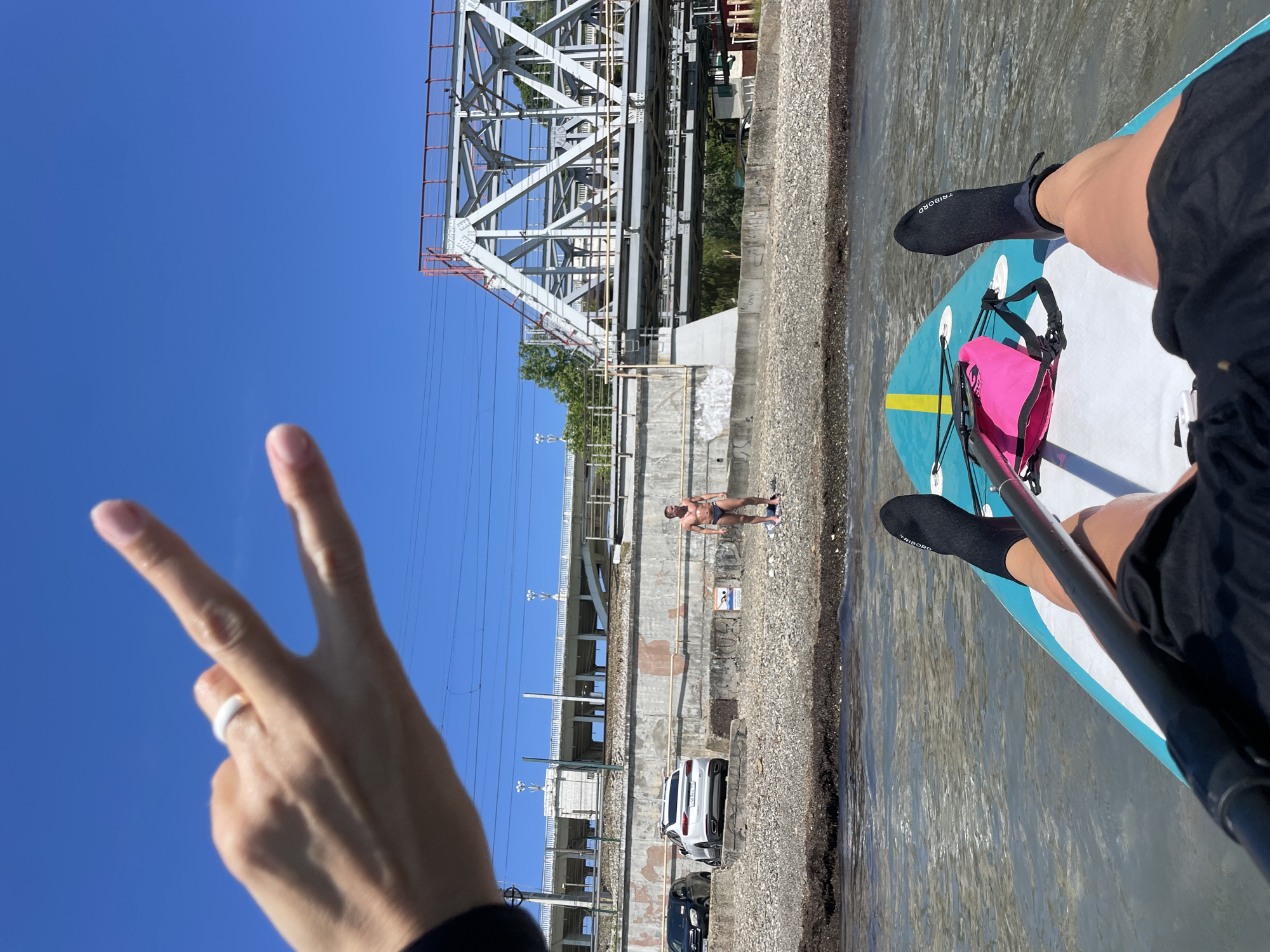 The image captures a vibrant, sunny day scene from the perspective of someone paddleboarding on a body of water. The vantage point looks towards a shore and extensive industrial or urban infrastructure.
In the foreground, a hand extends from the left, forming a "peace" sign against the clear blue sky. A white ring is visible on one of the fingers. On the right, the legs and feet of another person (likely the paddleboarder taking the photo) are visible, clad in black water socks, one of which clearly displays "TRIBORD" in white text. A bright pink dry bag is secured to the front of the light blue and white paddleboard, which partially fills the lower right of the frame. The shaft of a paddle is also seen.
The water, reflecting the clear sky, appears calm. A rocky or gravelly shoreline borders the water on the left, behind which rises a concrete embankment. On this shore, a man wearing dark swim trunks stands facing the water. Further back, two cars are parked: a white SUV followed by a darker sedan.
Dominating the midground and background is a large, grey metal truss bridge, likely a railway bridge, with red safety railings. Beneath and around the bridge, concrete structures, power lines, and some greenery are visible, indicating an urban or industrial setting. The clear, bright blue sky suggests excellent weather conditions for water activities. A small blue sign is visible on the concrete embankment near the man on shore, though its specific text is illegible. The overall impression is one of leisure and outdoor activity on a bright day in an urban waterside environment, consistent with a location like Sochi.