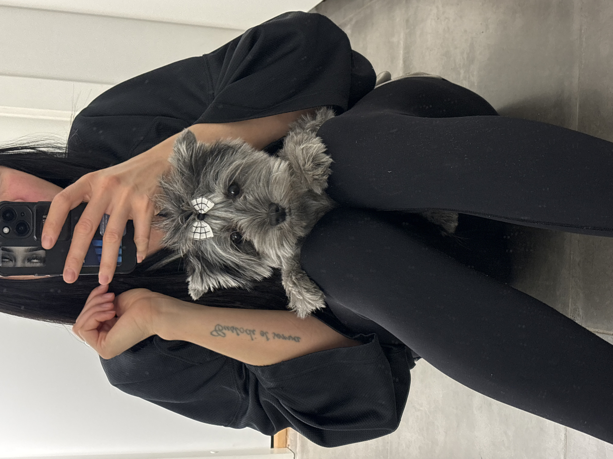 A person, visible from the forehead down and holding a black cell phone with an eye sticker on it, is taking a selfie with a small grey dog that has white bows on its head. The person is wearing black leggings and a black top. The dog is sitting in the person's lap. A tattoo is visible on the person's left forearm. The background appears to be a wall, possibly in a bathroom. The image is rotated counter-clockwise. There is no information to determine if the photo was taken in the city of Chaikovsky, Russia.