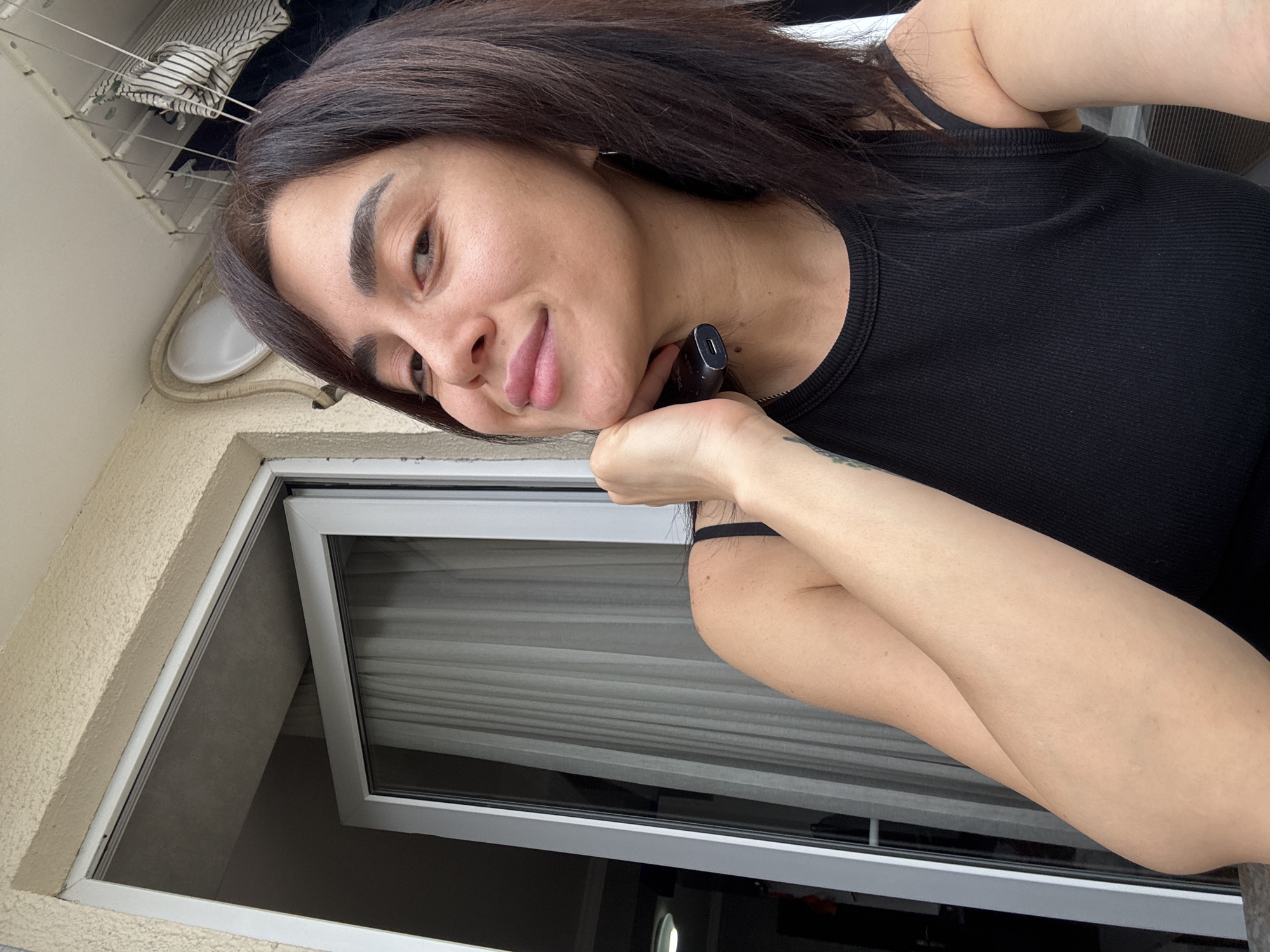 A woman is taking a selfie on a balcony in Tbilisi, Georgia. She has dark hair and is wearing a black tank top. She is holding a black electronic device to her chin. The balcony has a railing and a view of a building with a window and curtains. There is also a clothesline with clothes hanging on it in the background. The woman is smiling and looking at the camera.
