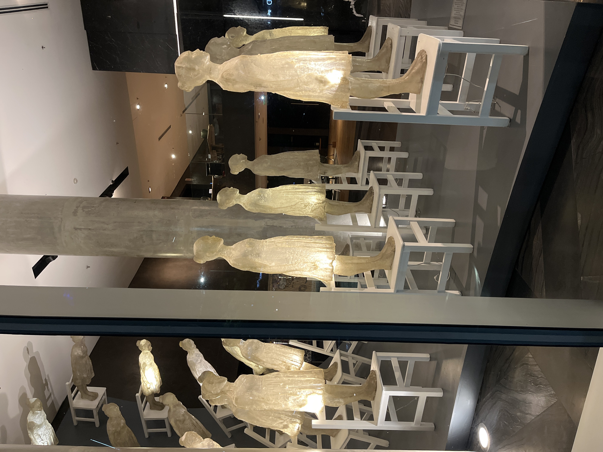 The image depicts an art installation in a display window, likely in Buenos Aires, Argentina. Several translucent, human-like figures, possibly made of resin or glass, stand on white chairs arranged in rows. The figures appear to be female and are positioned as if they are sitting. The display window reflects the interior of a building, including a large cylindrical pillar and other architectural elements, indicating an indoor location. The lighting within the display illuminates the figures and chairs, creating a soft, ethereal glow. The overall impression is that of a contemporary art piece displayed in a public space.